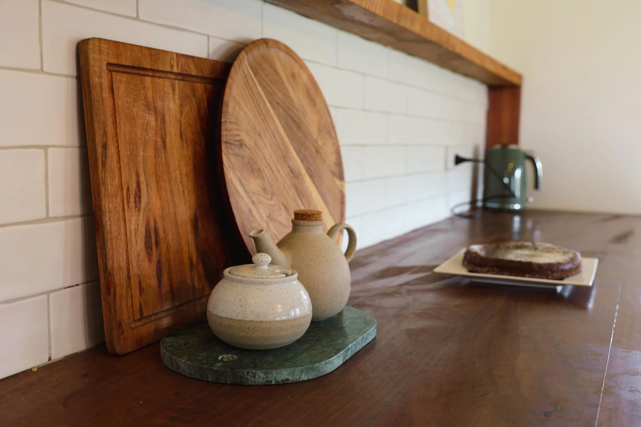 Countertop with cutting boards, ceramic teapot and sugar jar, a plate with meat, in a kitchen with a white brick wall and a green electric kettle.