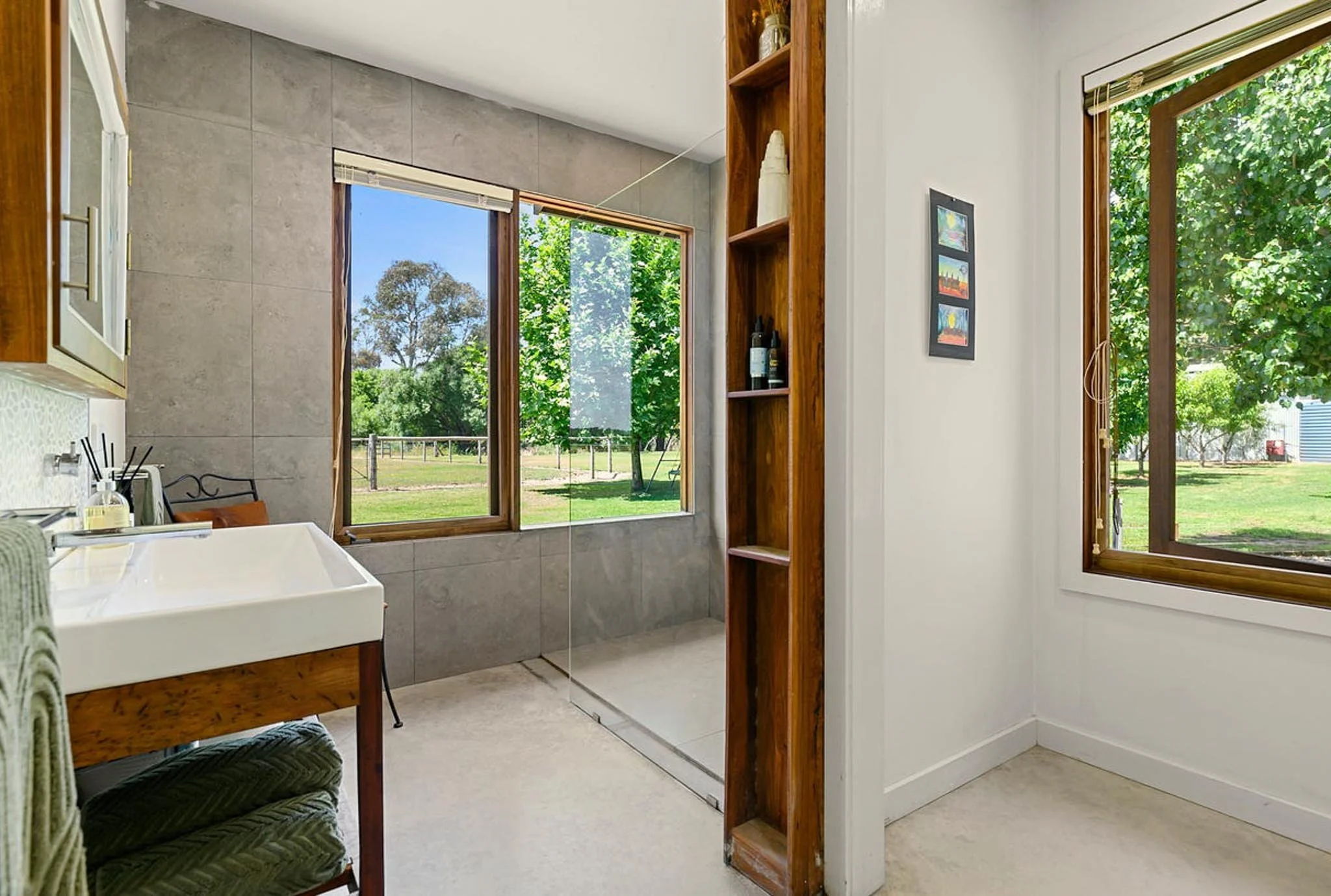 View of a bathroom with large windows showing green trees and an outdoor landscape, a white sink, and a wooden shelving unit.