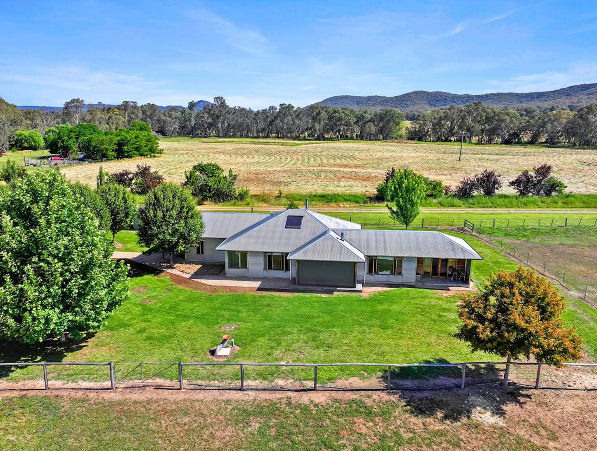 A house with a metal roof situated on a lush green lawn surrounded by trees, with rolling fields and hills in the background under a blue sky.