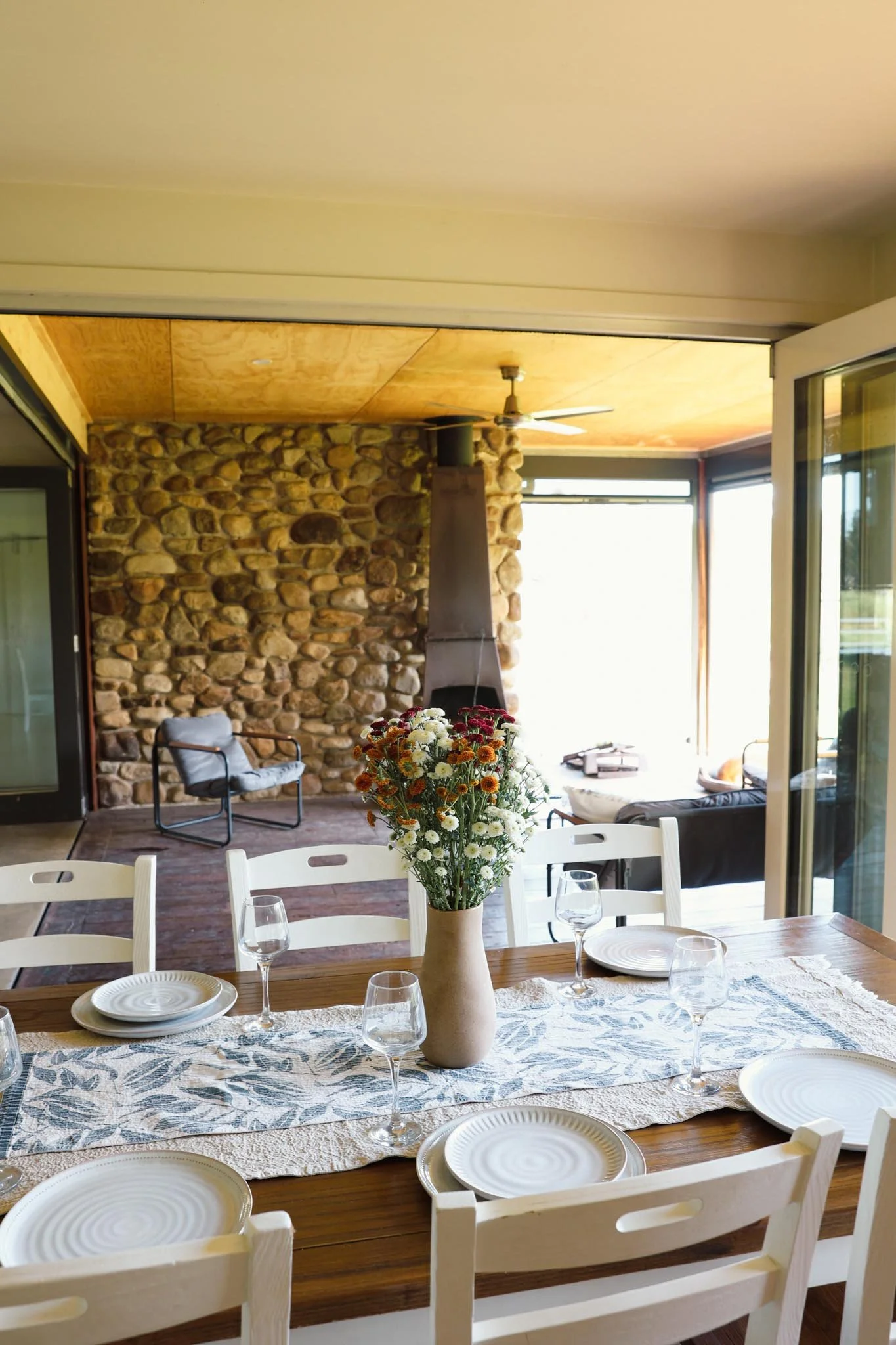 A dining table set with plates, glasses, and a vase of wildflowers in a bright dining area with large windows and a stone fireplace in the background.