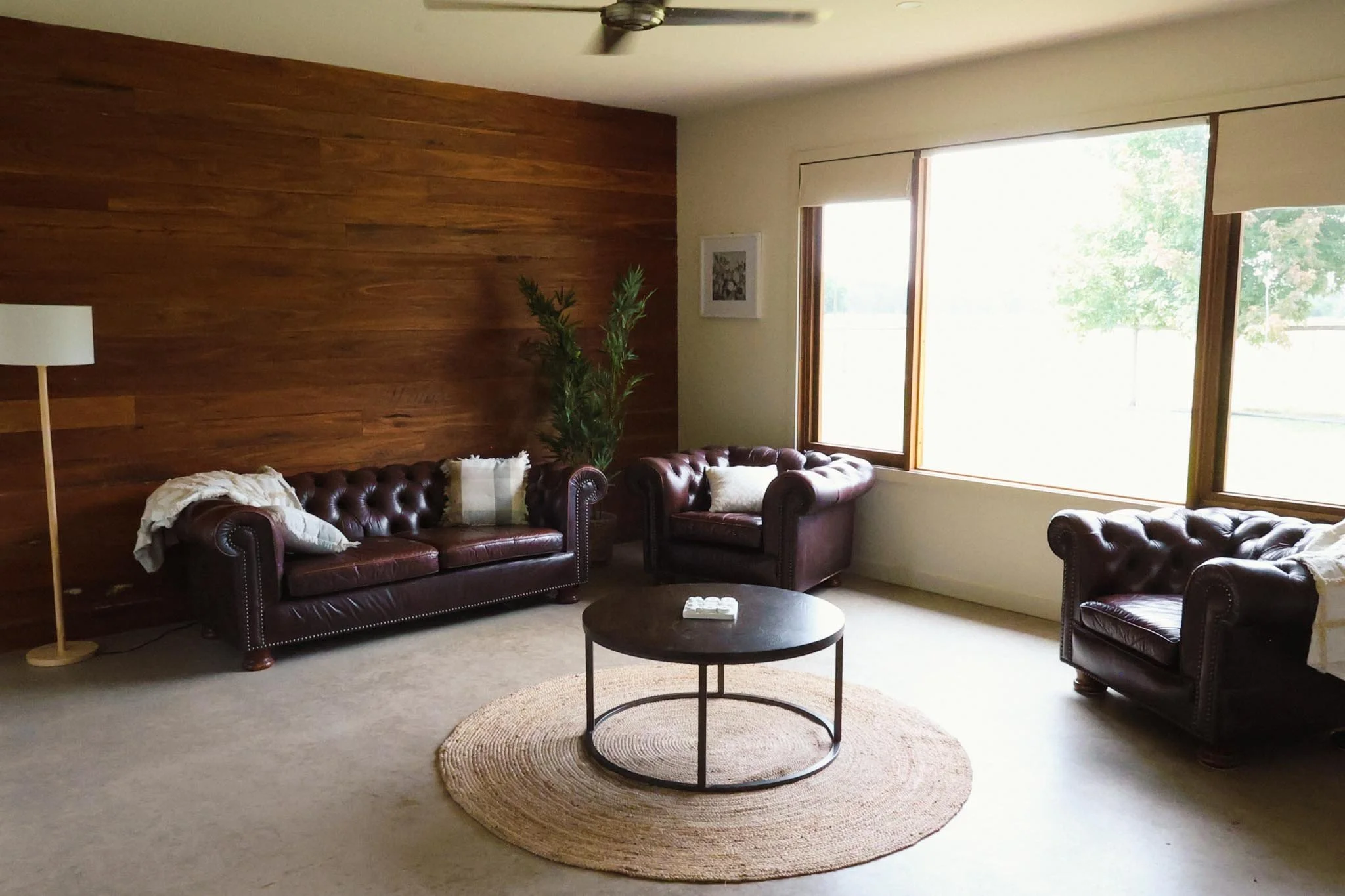 Living room with three leather sofas, a round coffee table, a tall floor lamp, a potted plant, and a large window letting in natural light. The wall behind the sofas is wood-paneled.