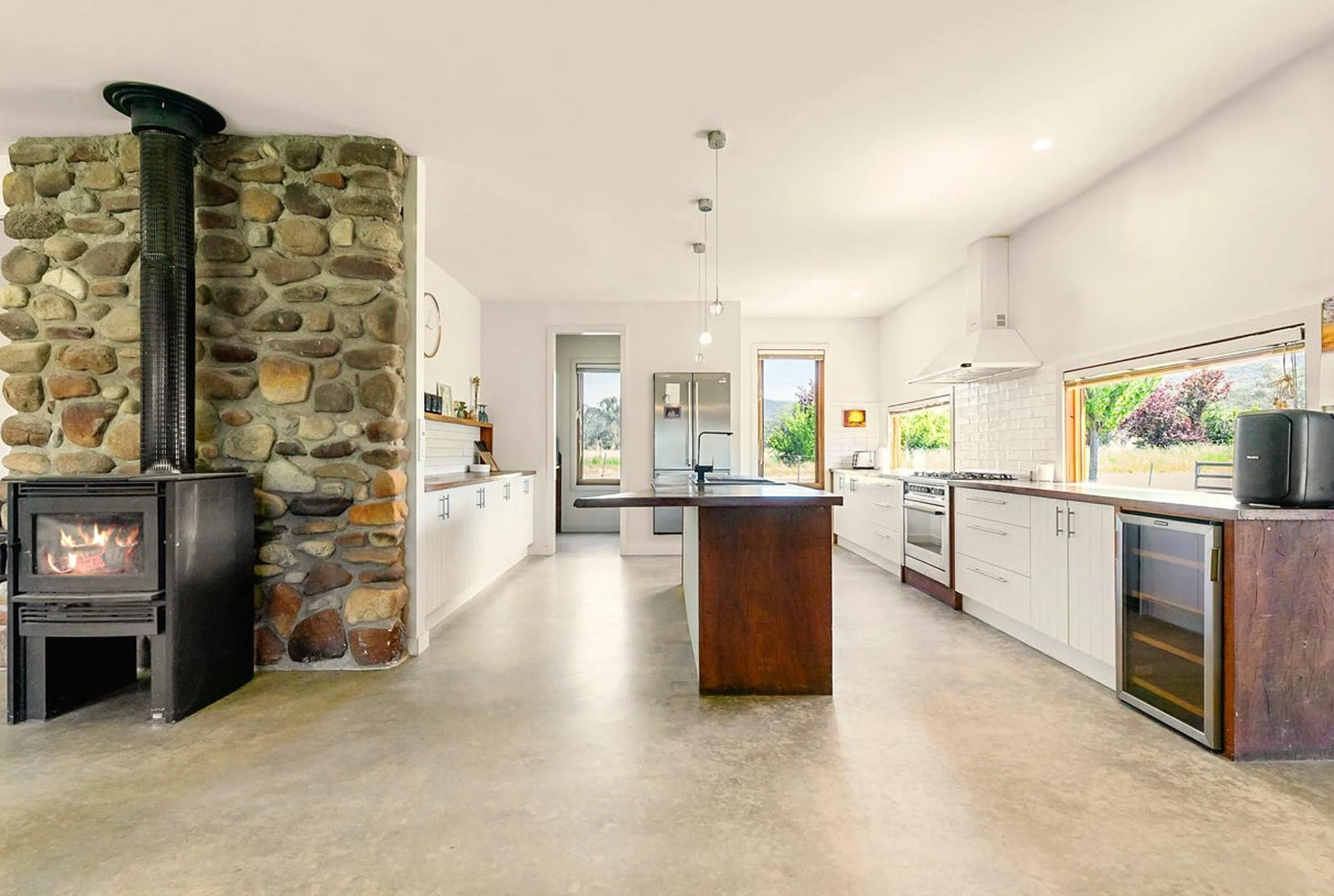 Spacious modern kitchen with white cabinets, a wooden kitchen island, stainless steel appliances, large windows with outdoor views, and a stone fireplace with a wood stove.