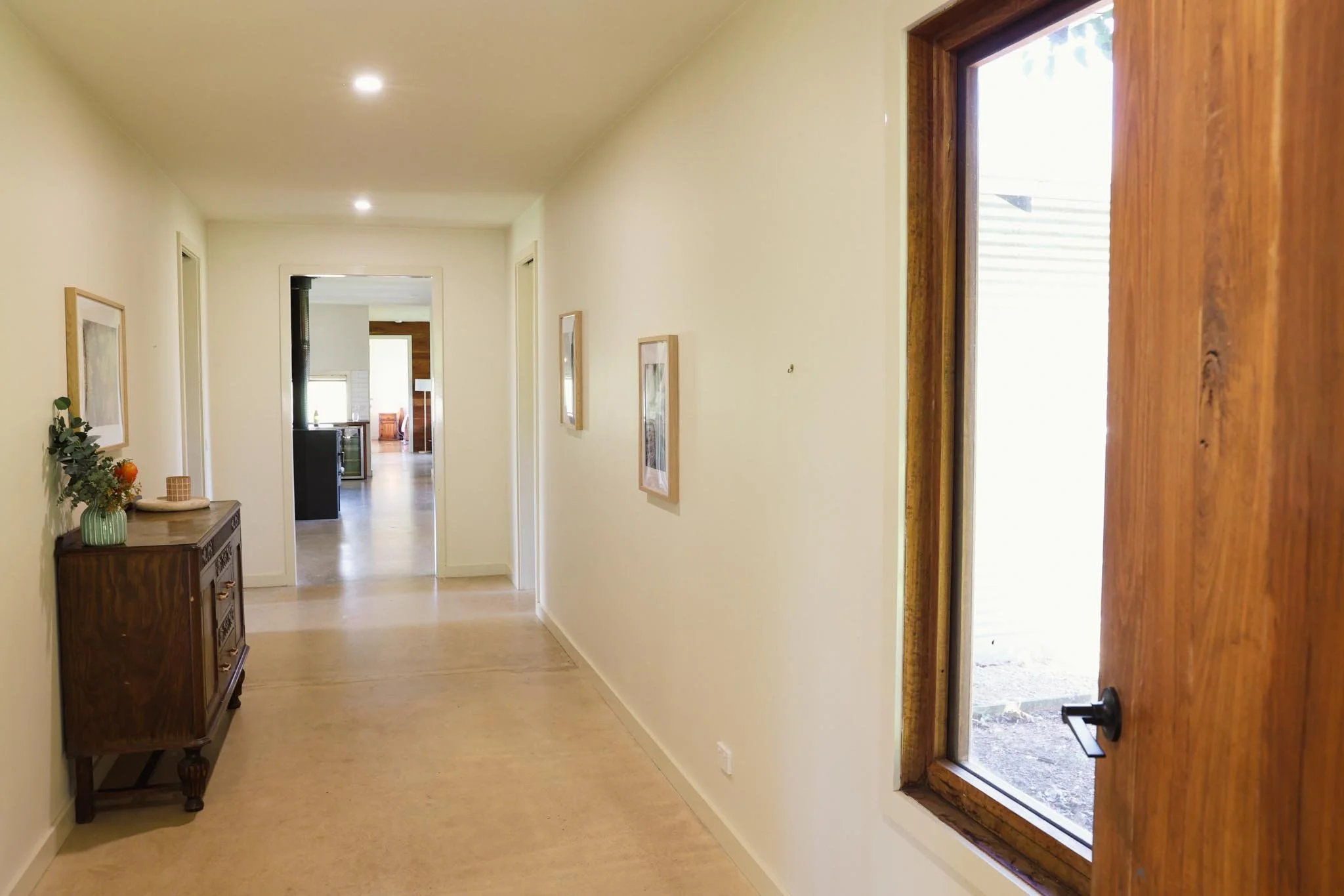 An interior hallway with cream-colored walls, wooden flooring, and framed artwork. On the left, there is a dark wooden console table with decorative items, including a vase with flowers. On the right, there is a window with a wooden frame, through wh