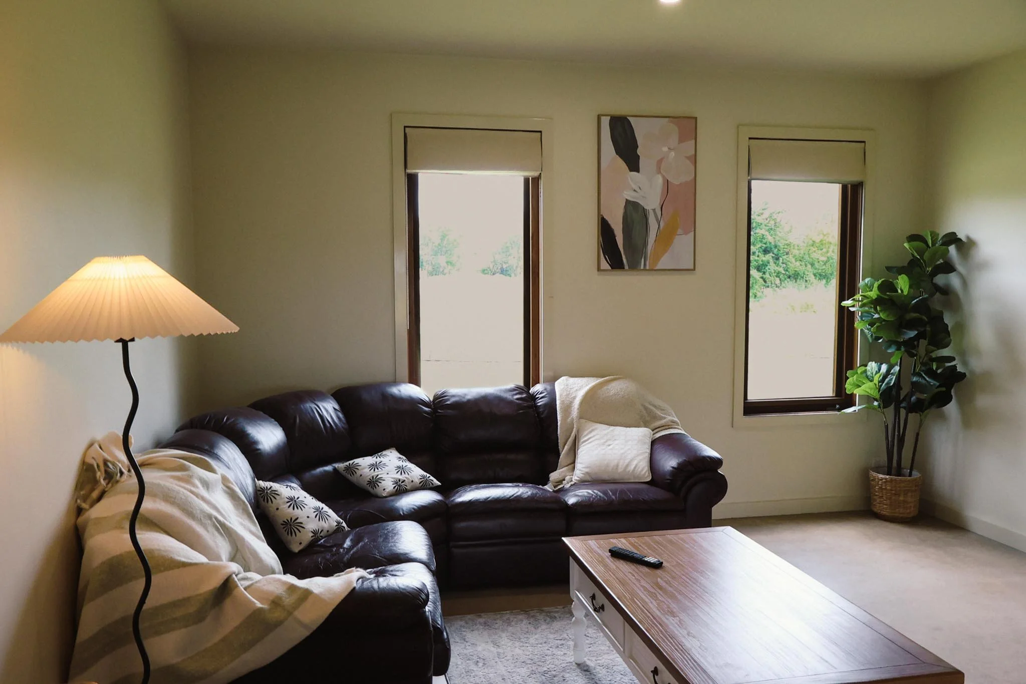 Living room with a black leather sectional sofa, a wooden coffee table, a tall floor lamp, a framed abstract floral painting, two windows with white blinds, a potted plant, and some cushions and blankets.