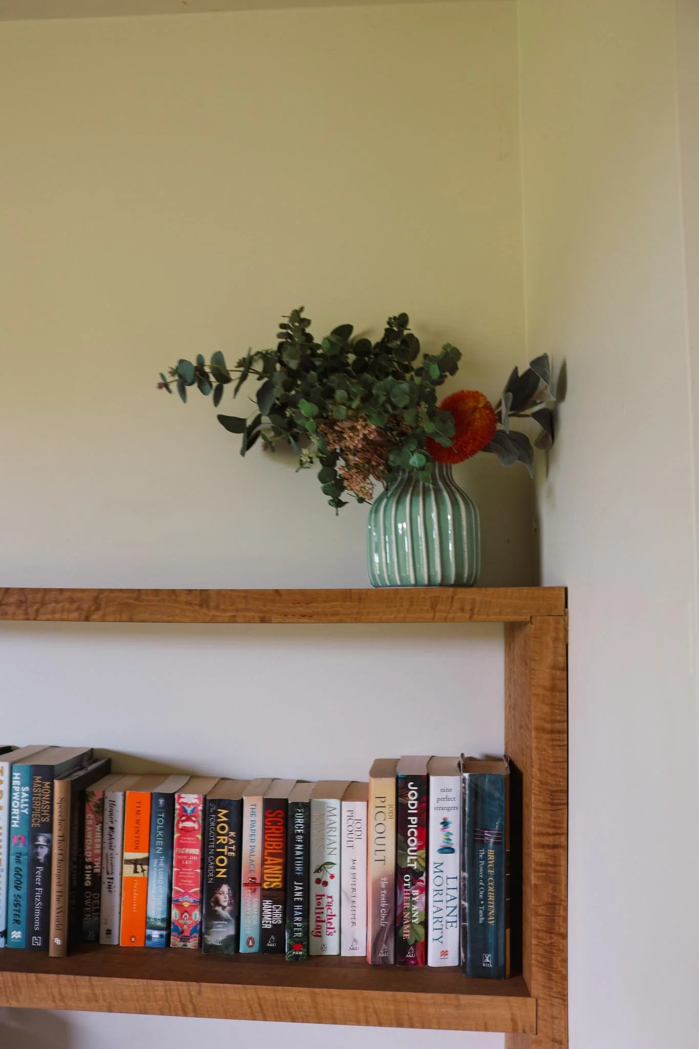 A green striped vase with flowers on a wooden shelf, and books on a lower shelf.