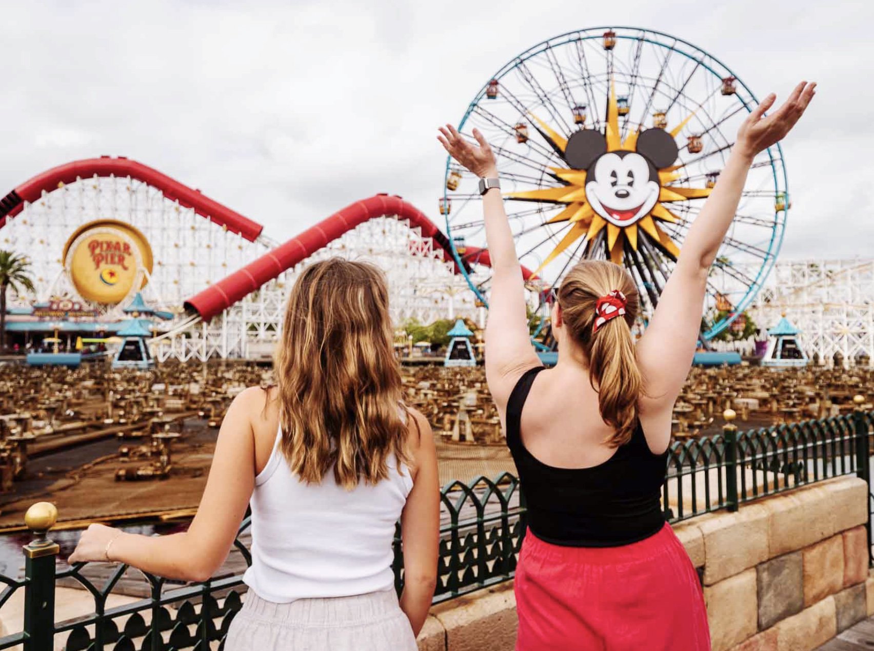 Two women standing by the fence at an amusement park, looking at a large Mickey Mouse Ferris wheel and a roller coaster at Pixar Pier in the background, with cloudy skies overhead.