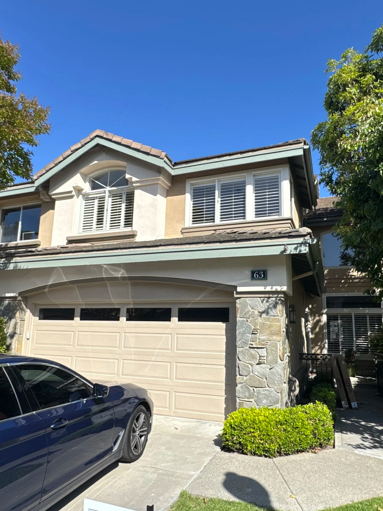 Front view of a two-story house with a beige garage door, stone accents on the lower part, off-white walls, multiple windows with white shutters, and a black car parked in the driveway. There is a small green bush near the sidewalk and trees on the sides.