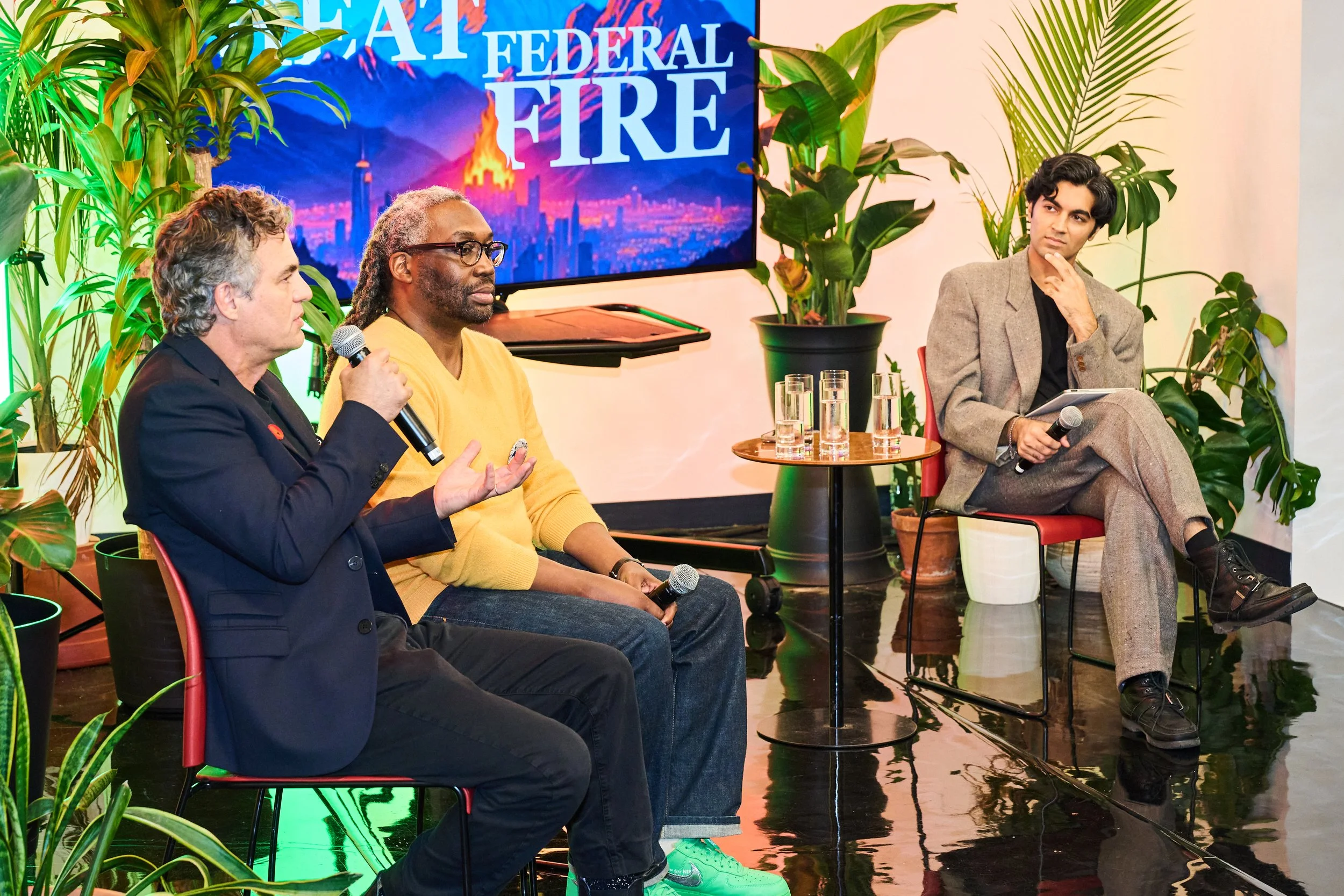 A panel discussion with three men seated on stage, surrounded by plants, with a screen displaying the words 'FIRE' behind them.