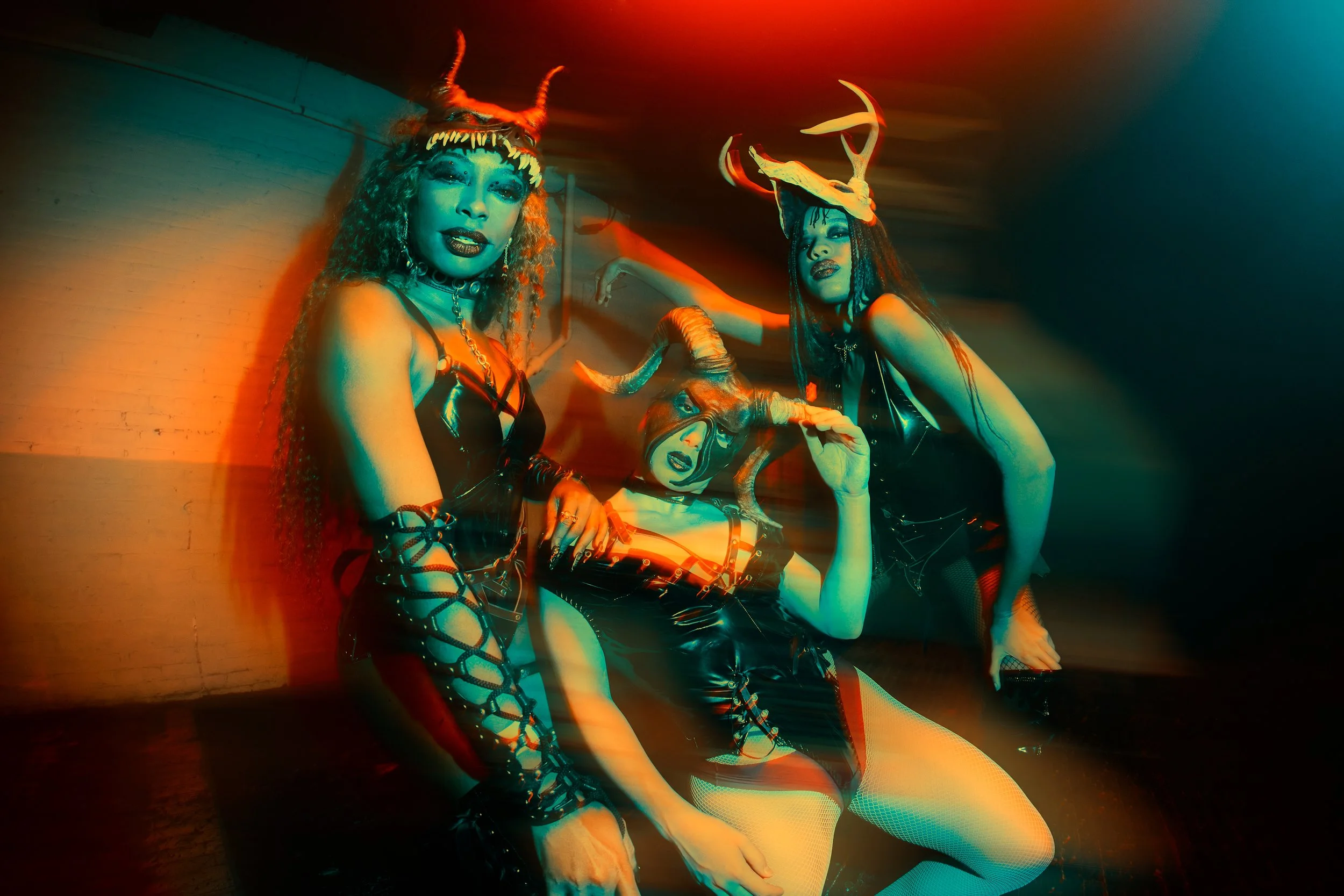 Three women in black leather outfits with horned and demon-themed costumes at a party, posing dramatically with colored lighting.