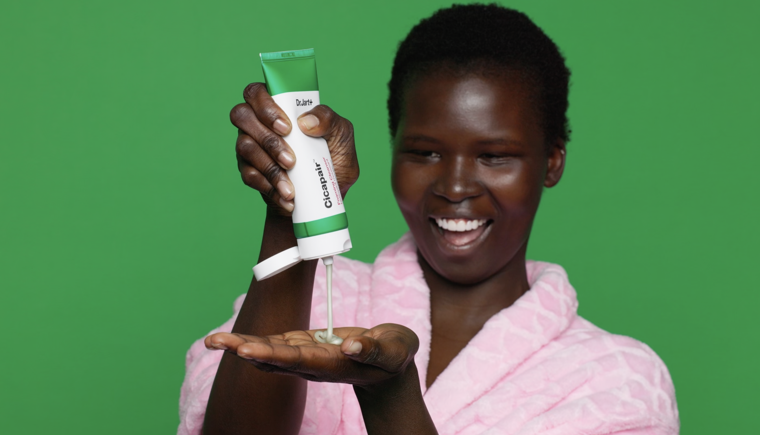 A smiling woman in a pink bathrobe holds a tube of Cicapair skincare cream, dispensing some onto her palm against a green background.