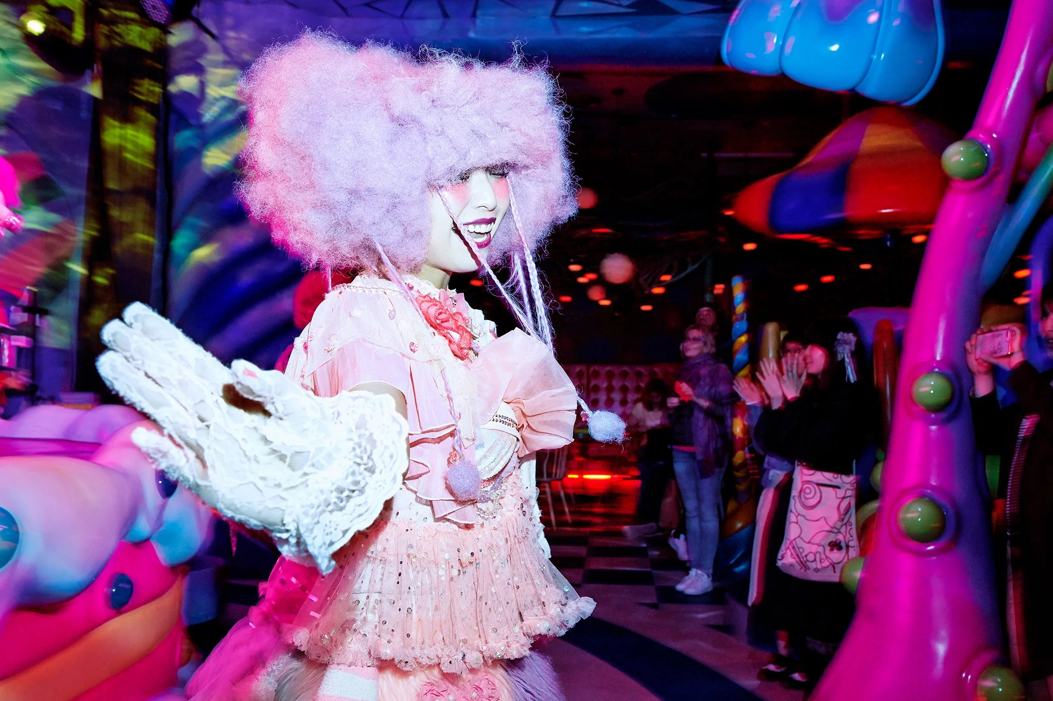 A person dressed as a clown with pink fluffy hair, white face makeup, and colorful clothing is playing a game at an amusement park or arcade. There are people in the background, with colorful lights and playful decorations around.