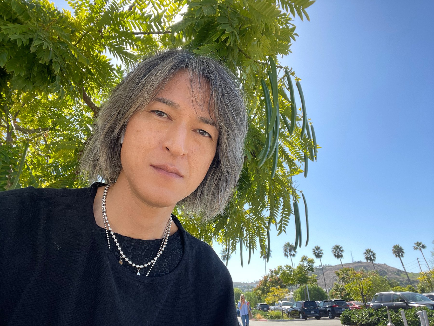 A person with shoulder-length gray hair, wearing a black shirt and layered necklaces, outdoors with a green leafy tree behind them, blue sky, and a parking lot with cars and palm trees in the background.