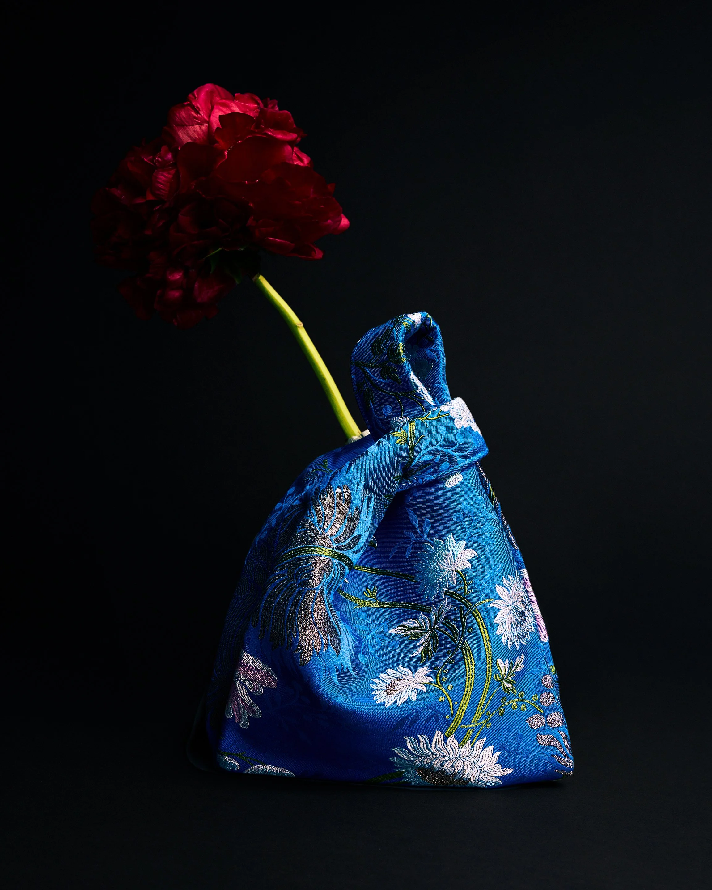 A red flower with a long green stem is placed inside a blue silk bag with floral embroidery, against a black background.