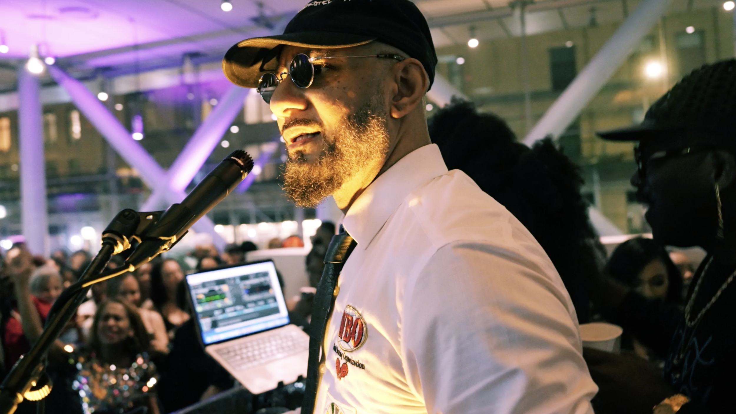 A DJ with sunglasses and a black cap, wearing a white shirt with patches, is performing at a lively event with a crowd in the background. There is DJ equipment and a laptop visible.