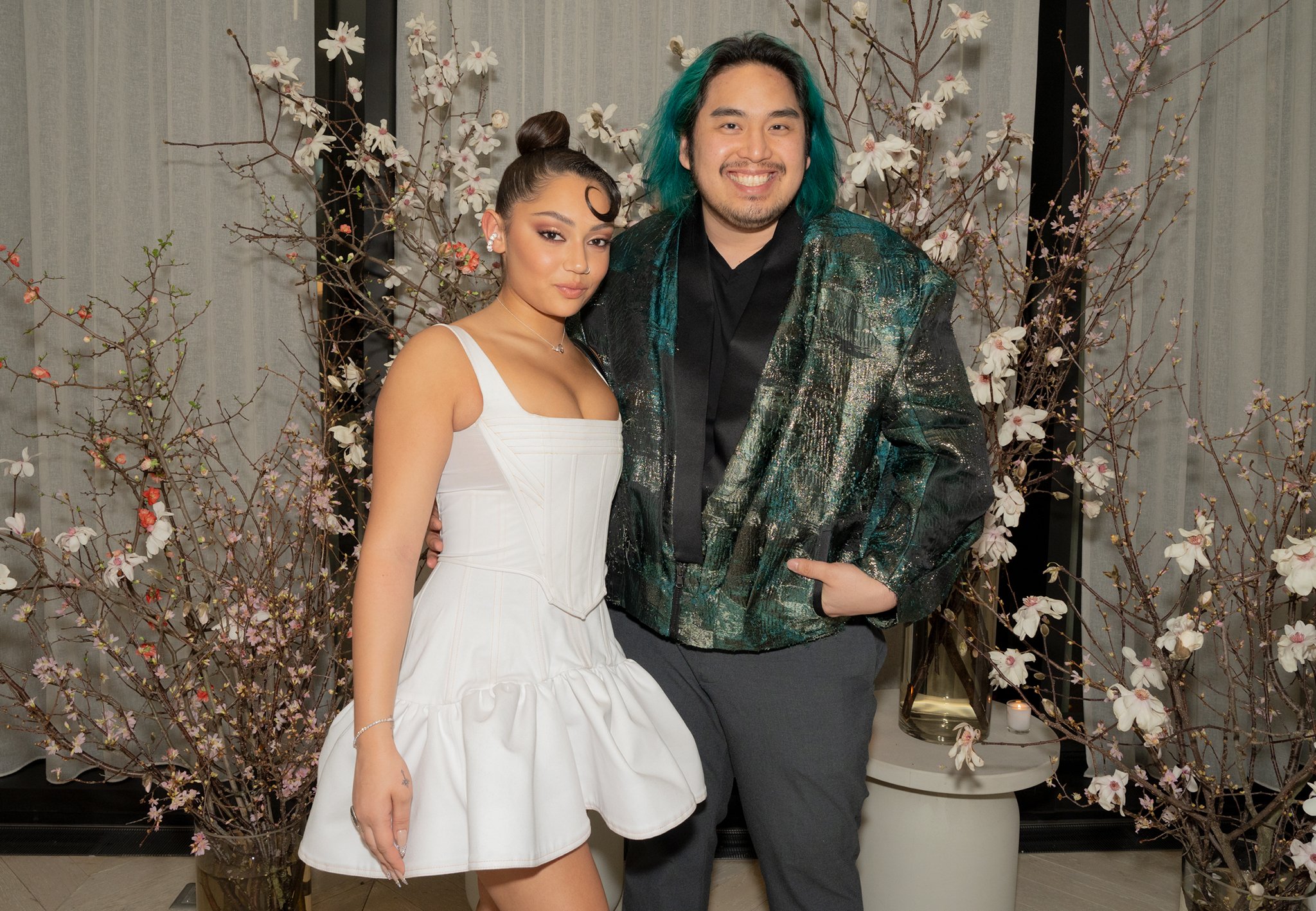 Two people, a woman in a white dress and a man in a shiny, patterned jacket, standing close together in front of floral arrangements and a curtain, smiling at the camera.