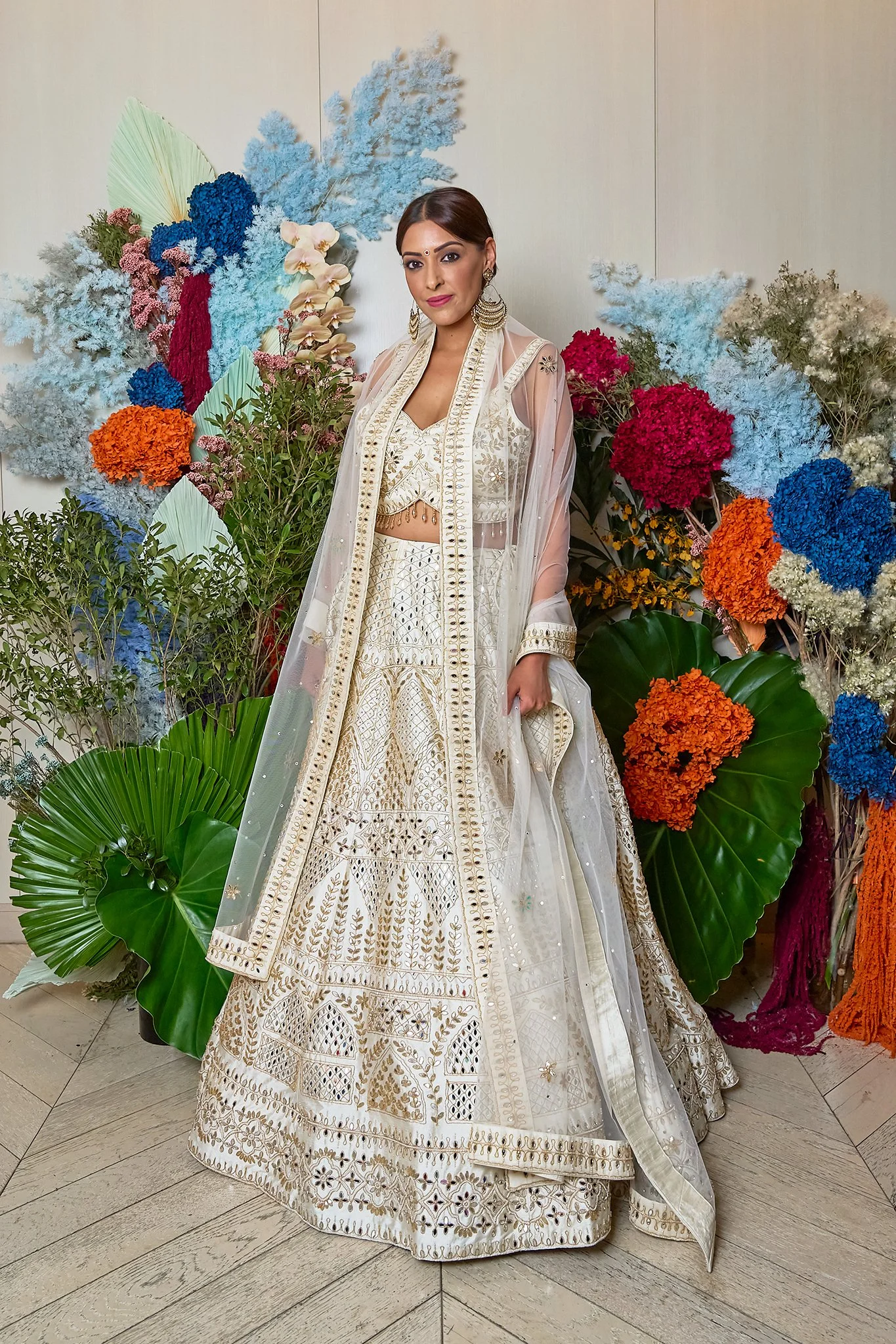 Woman in traditional Indian bridal attire standing in front of a colorful floral arrangement.