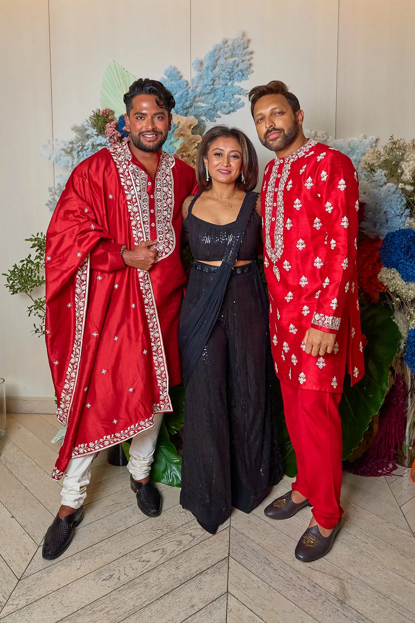 Three people in traditional Indian attire and a woman in black, standing in front of a colorful floral backdrop.