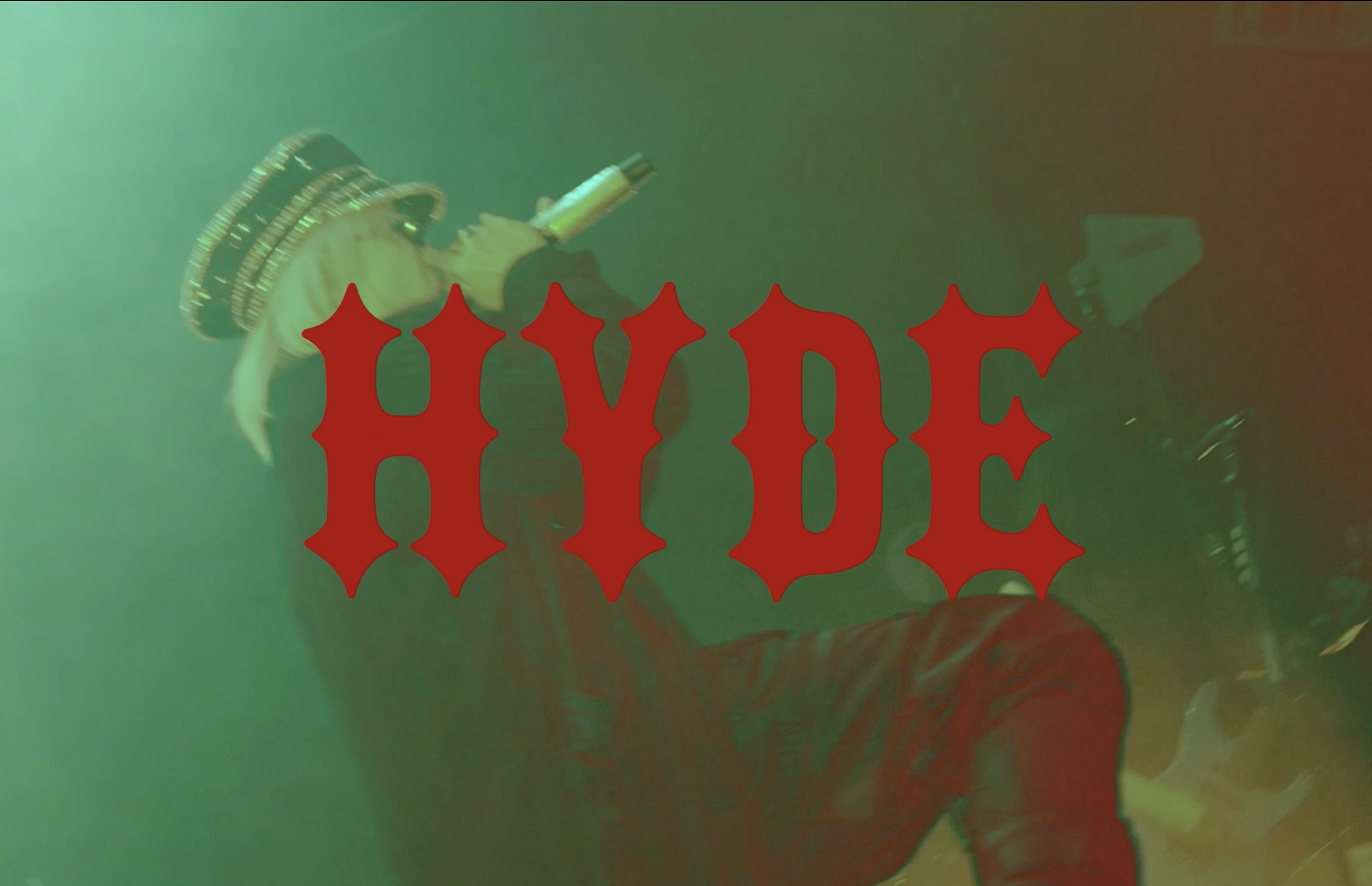 A person with long blond hair, wearing a leather jacket with spike shoulder pads, is holding a large gun and standing in front of a green and red background with the word 'HYPE' written in large red gothic font.