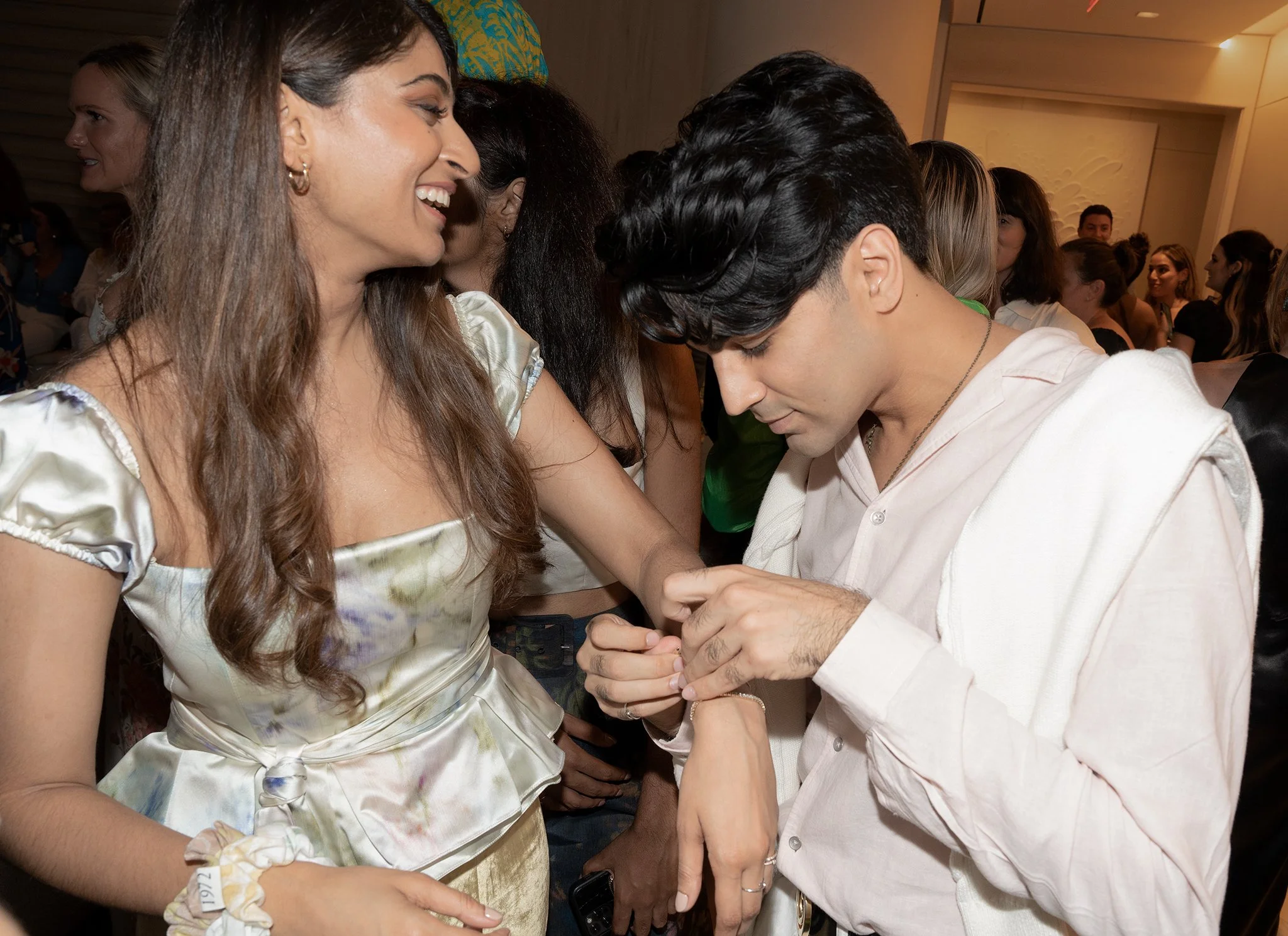 A woman with long brown hair smiling as a man with dark hair, dressed in a light-colored shirt, looks down and appears to be putting a ring on her finger in a crowded indoor setting.