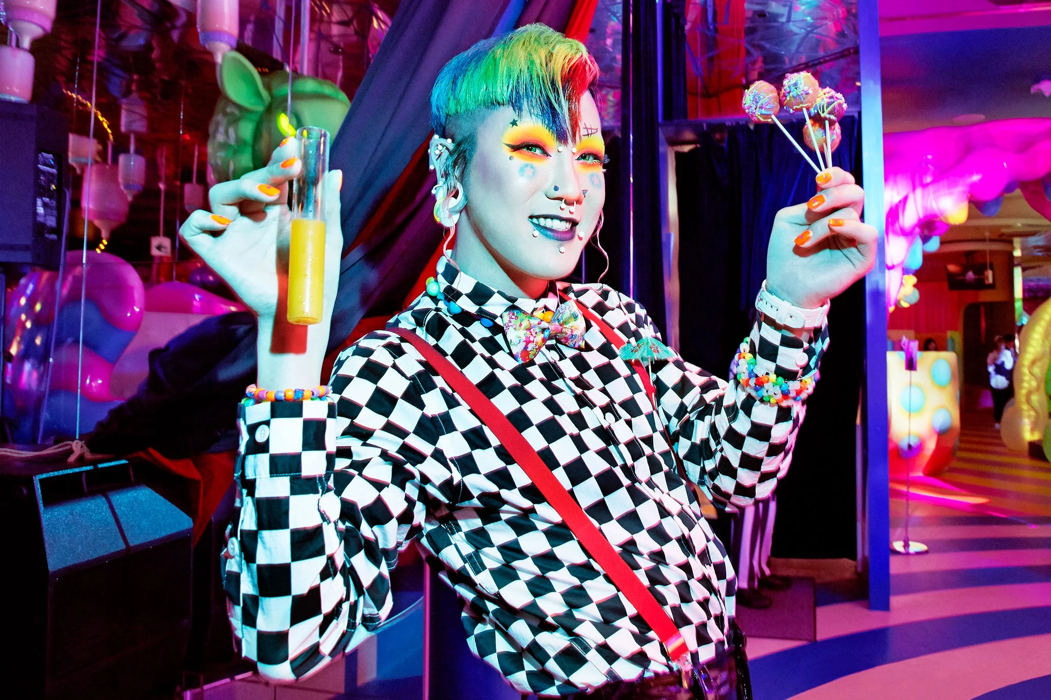 A woman with rainbow-colored hair and rainbow makeup poses at a colorful, neon-lit amusement park or carnival. She is wearing a black and white checkered shirt, colorful accessories, and holding marshmallow on a stick and a yellow drink.