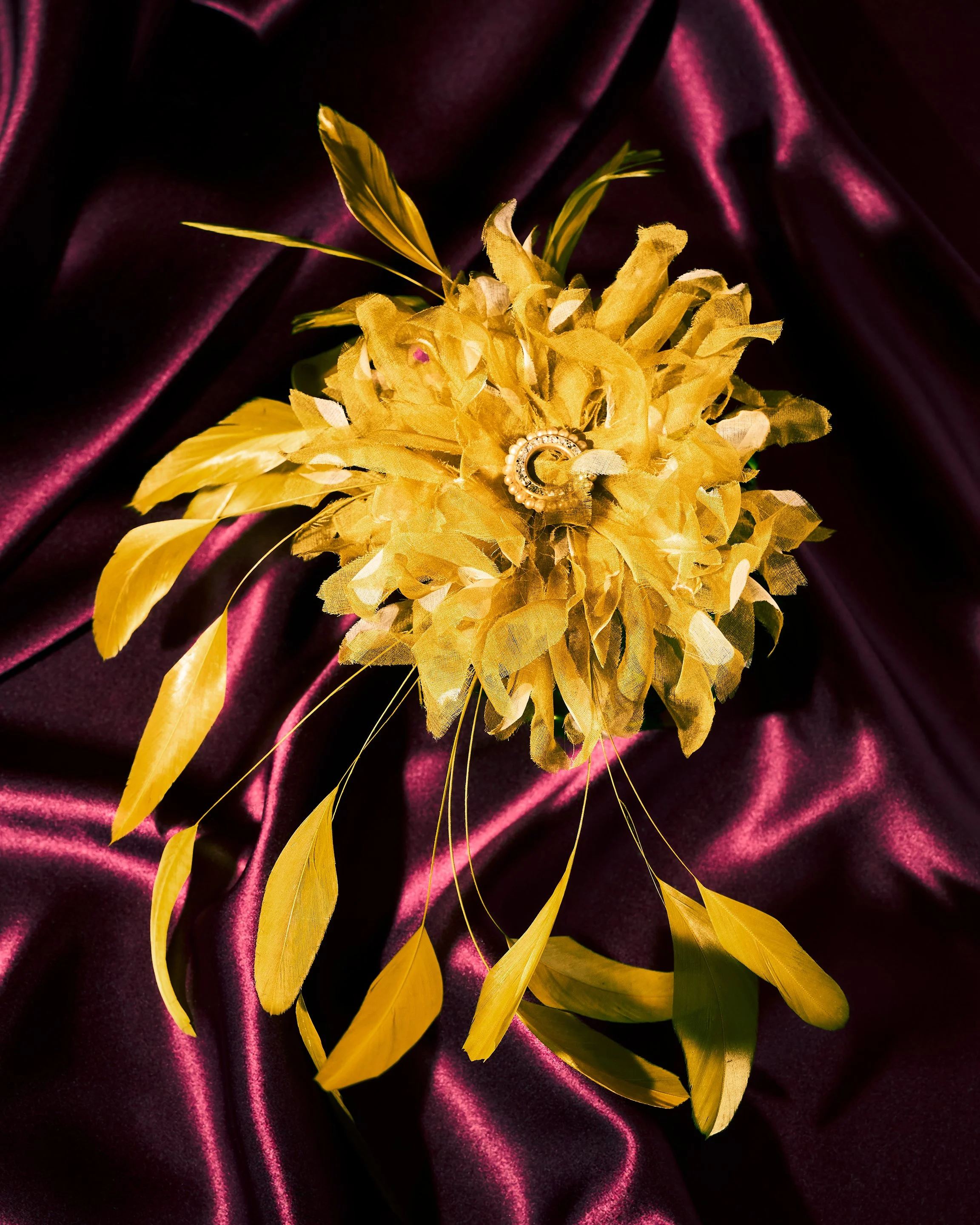 A yellow floral boutonniere with gold leaves and a pearl and gold brooch, on a dark purple satin fabric.