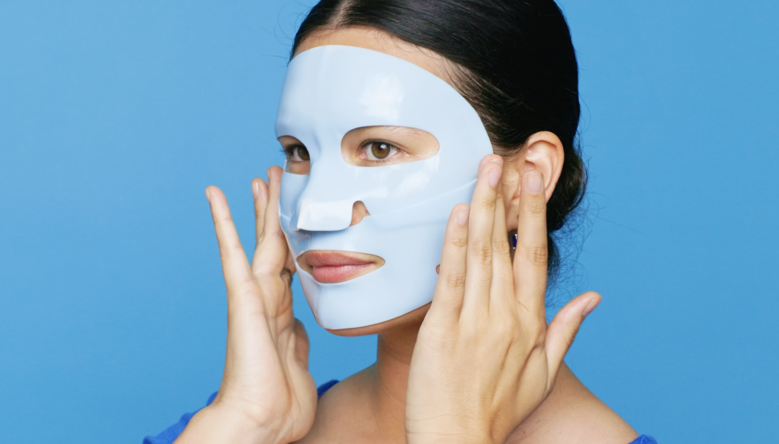 A woman with dark hair in a bun wearing a light blue facial mask against a blue background, touching her face with her hands.