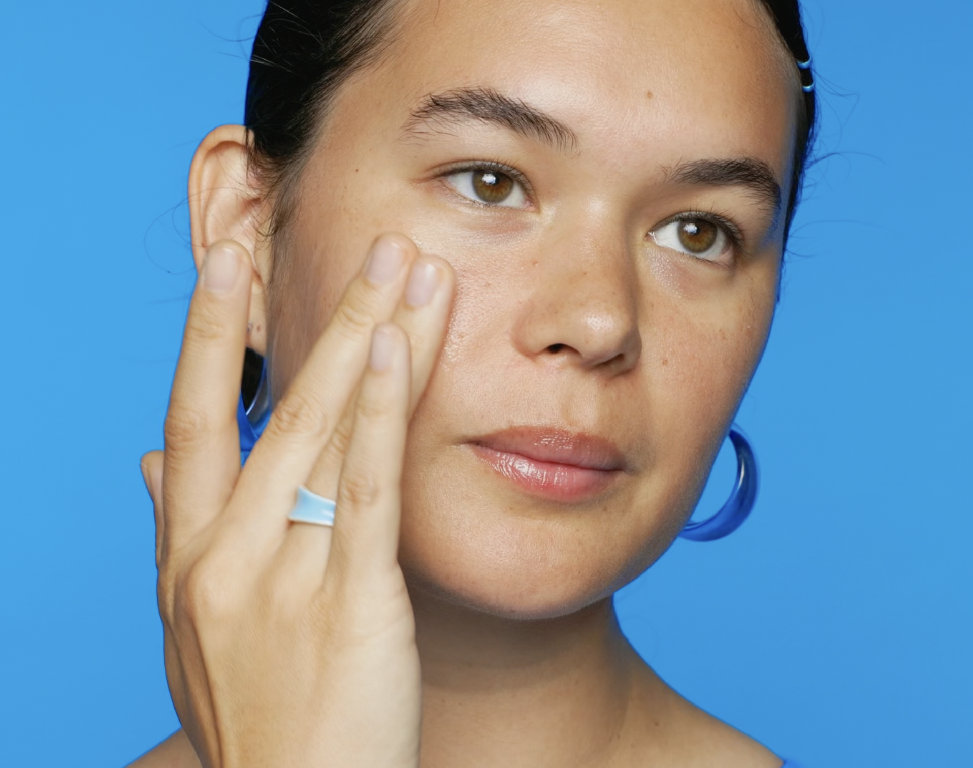 A woman with brown eyes and dark hair touching her cheek with her fingers, wearing a blue ring and hoop earrings, against a blue background.