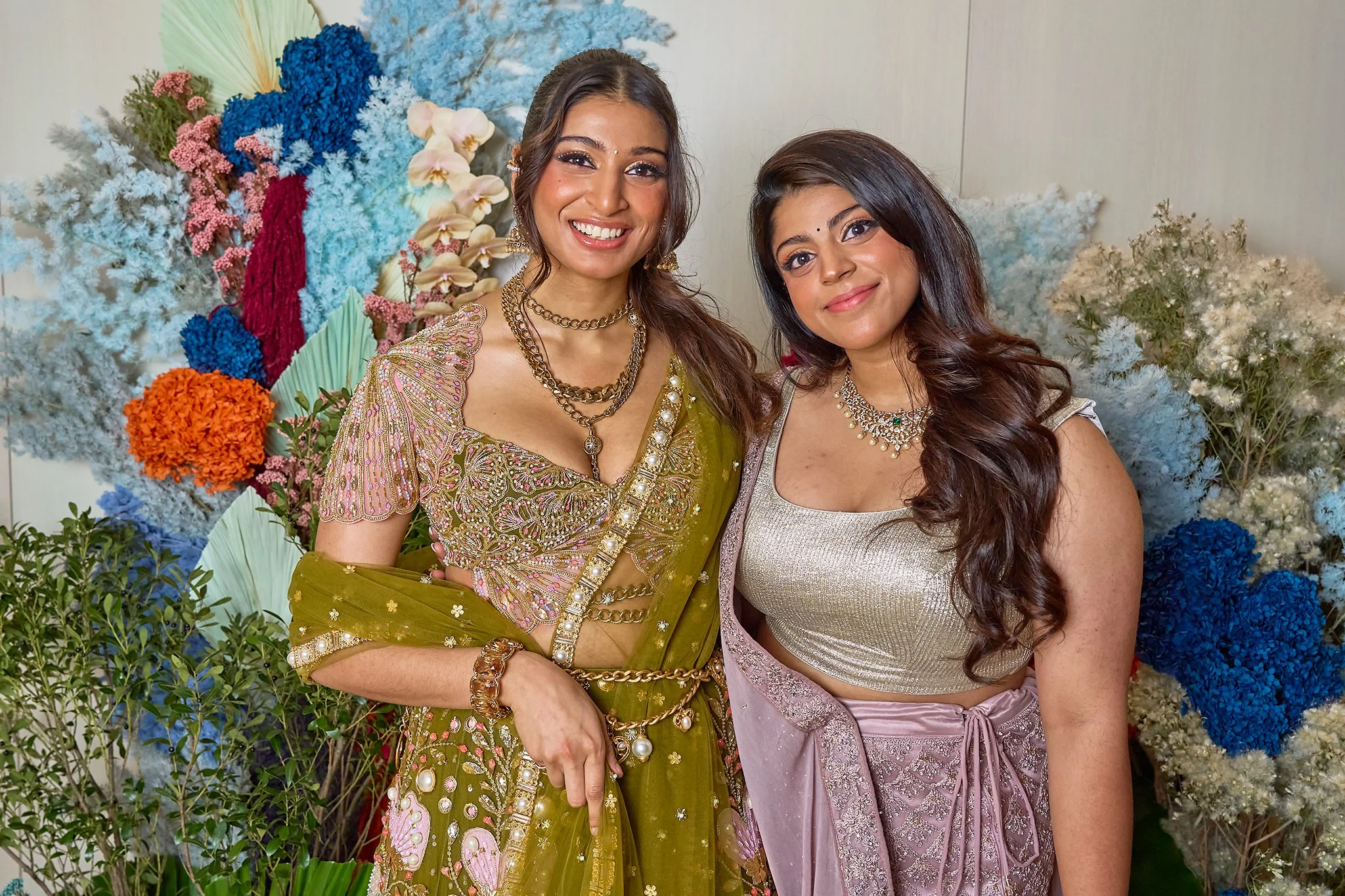 Two women in traditional Indian attire standing in front of colorful flowers, smiling at the camera.