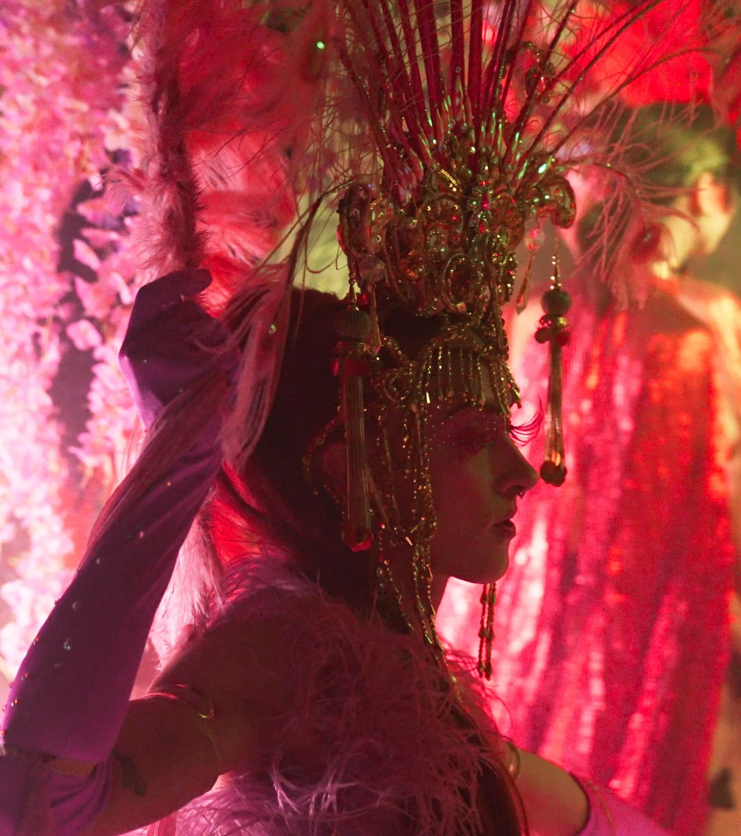 A person wearing an ornate headdress adorned with feathers, beads, and jewels, standing in dim pink lighting.