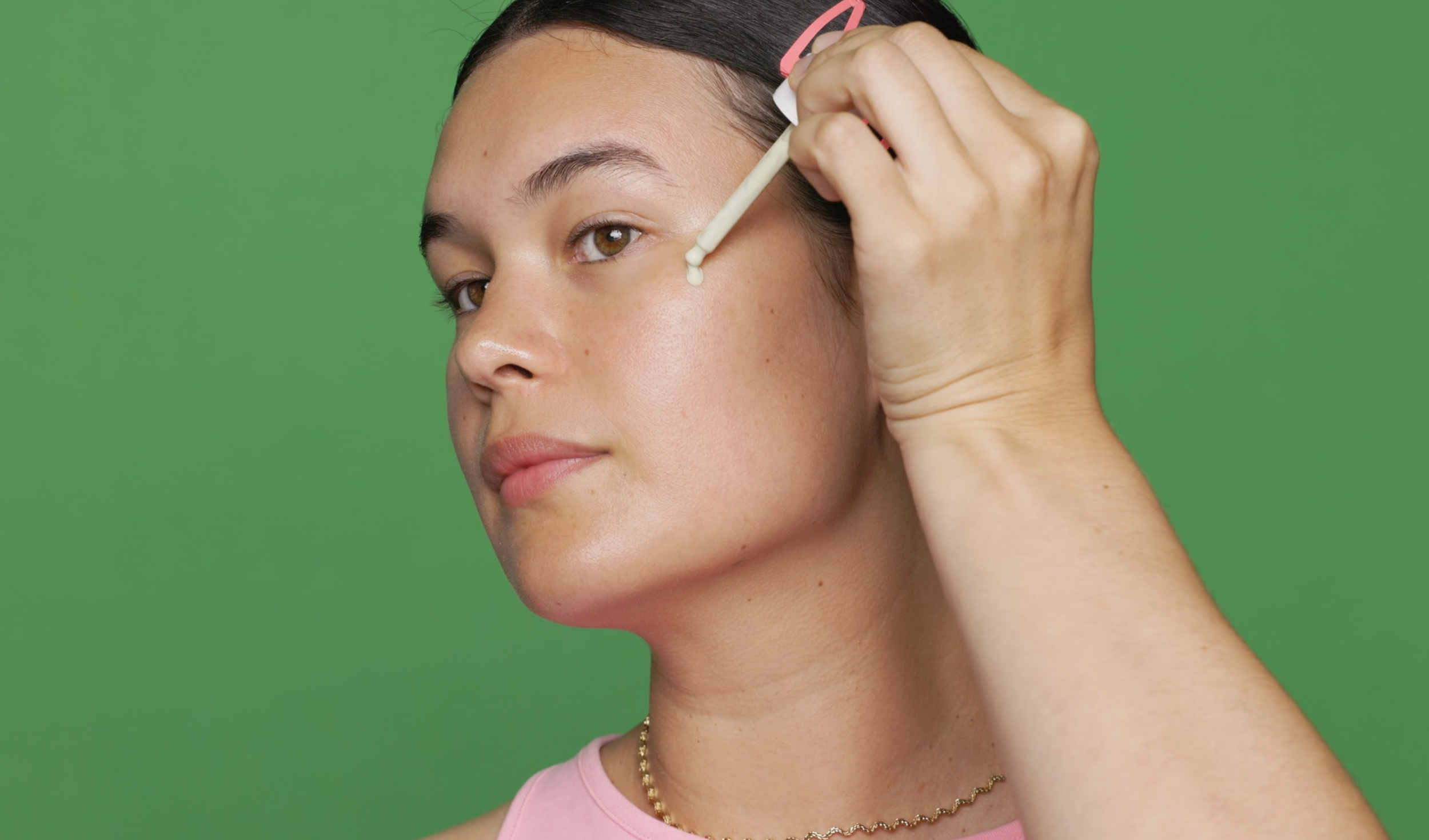 A woman applying a drop of skincare serum to her cheek using a dropper, against a green background.