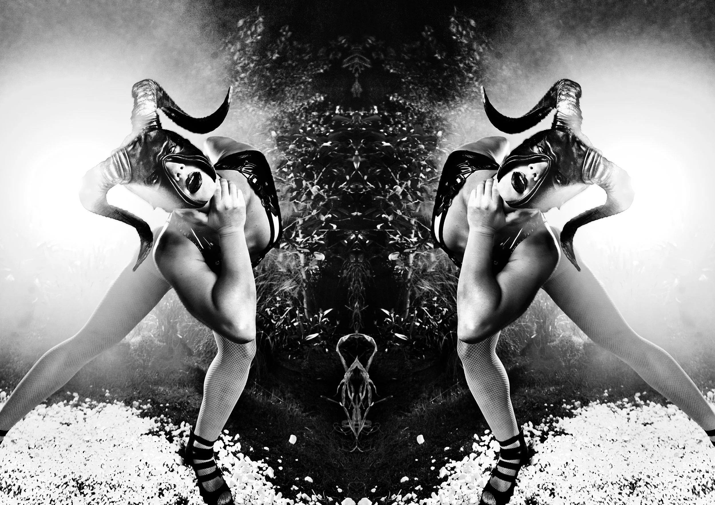 A black and white mirrored image of a woman in a provocative pose, wearing a mask with horns and latex clothing, standing outdoors on rocky ground with foliage in the background.