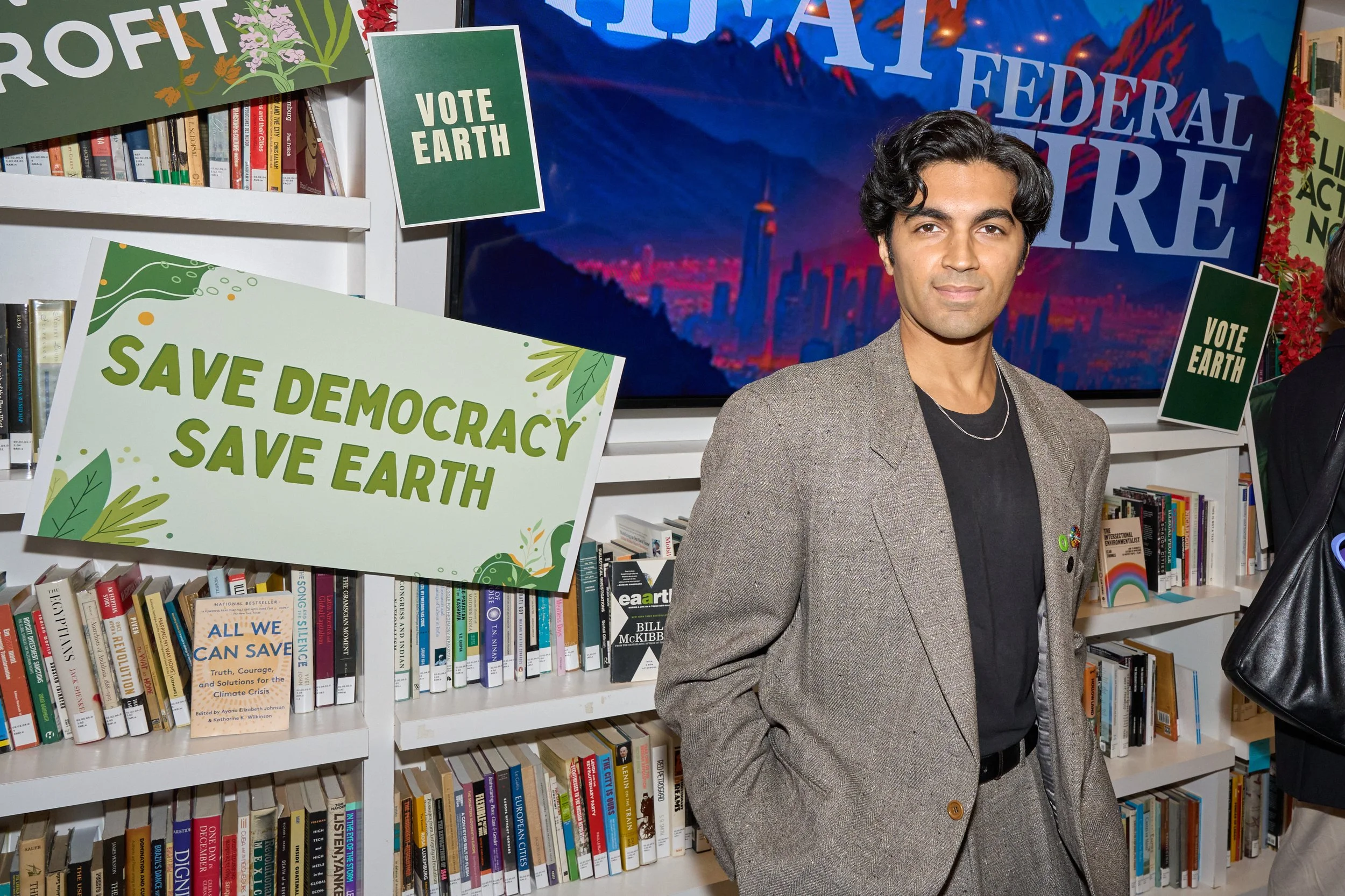 A young man in a suit standing in front of a bookshelf with political and environmental signs, including 'Save Democracy Save Earth' and 'Vote Earth', at a public event supporting environmental causes.