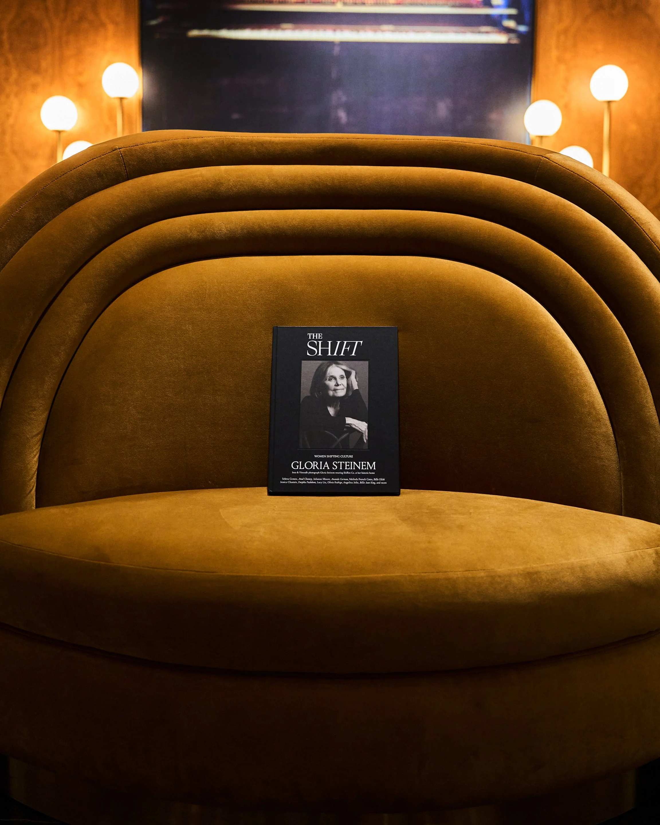 A black and white magazine titled 'The SHIFT' featuring Gloria Steinem on the cover, placed on a brown upholstered semicircular seating in a warmly lit room with wall lamps.