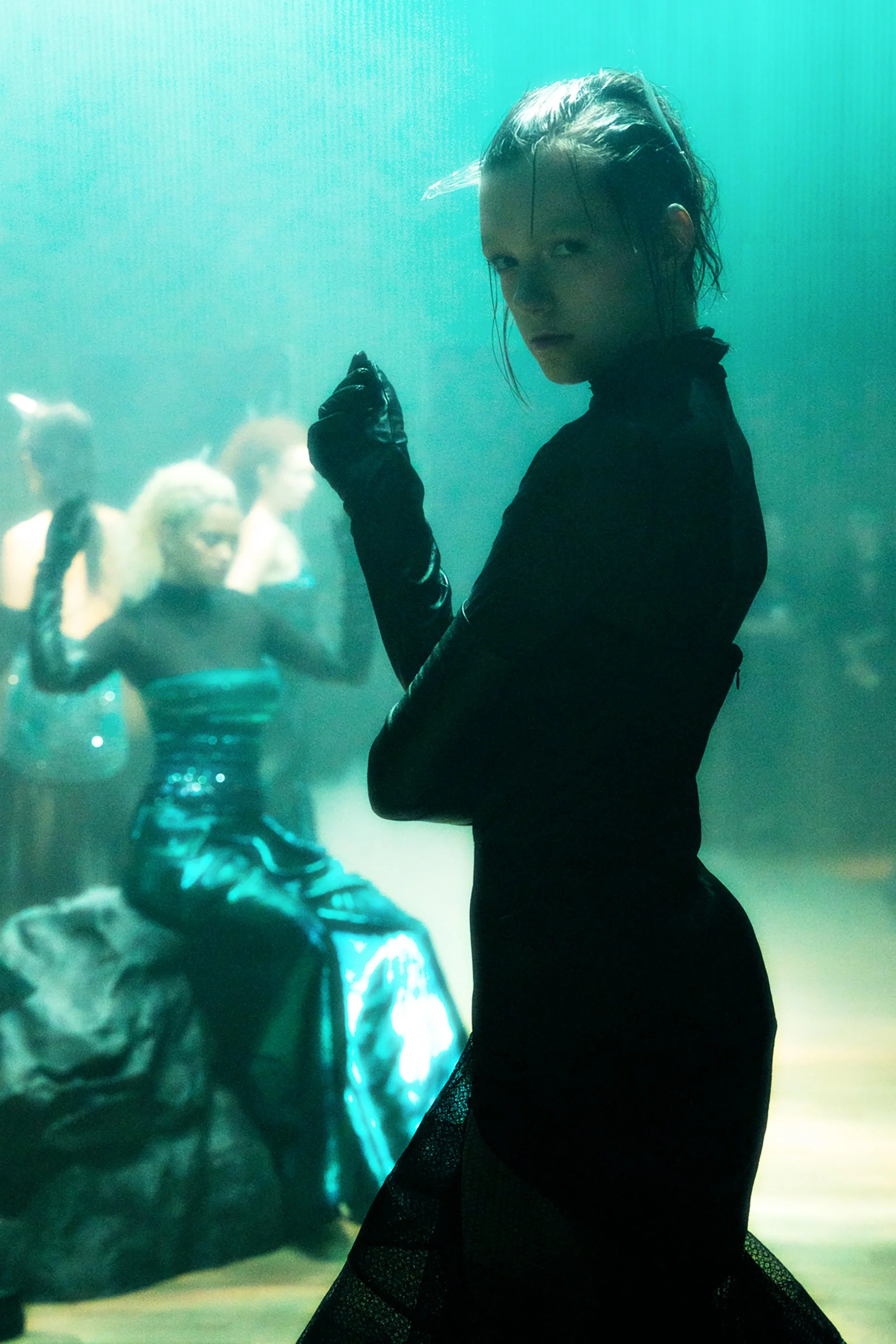 A woman in black with wet hair poses in front of an underwater scene with a statuesque figure and a tiger sculpture.