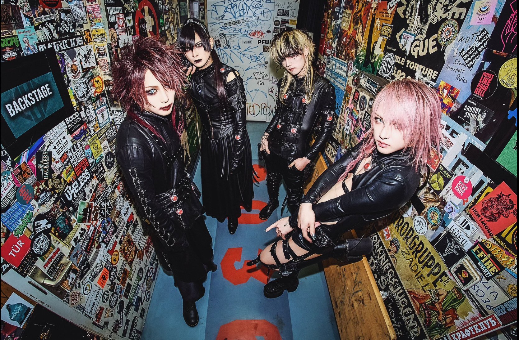 Four individuals dressed in black leather outfits with edgy hairstyles, standing in a narrow corridor covered with stickers and graffiti on the walls.