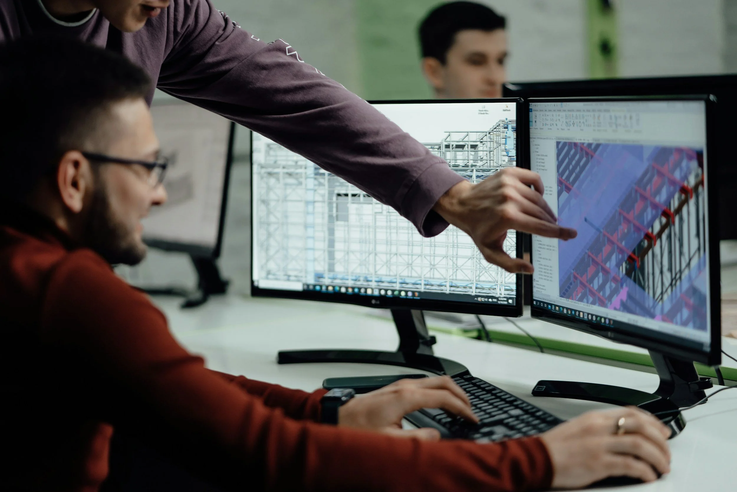 Two men working on computer screens displaying architectural and structural design plans, with one man pointing at the screens and the other typing on a keyboard.