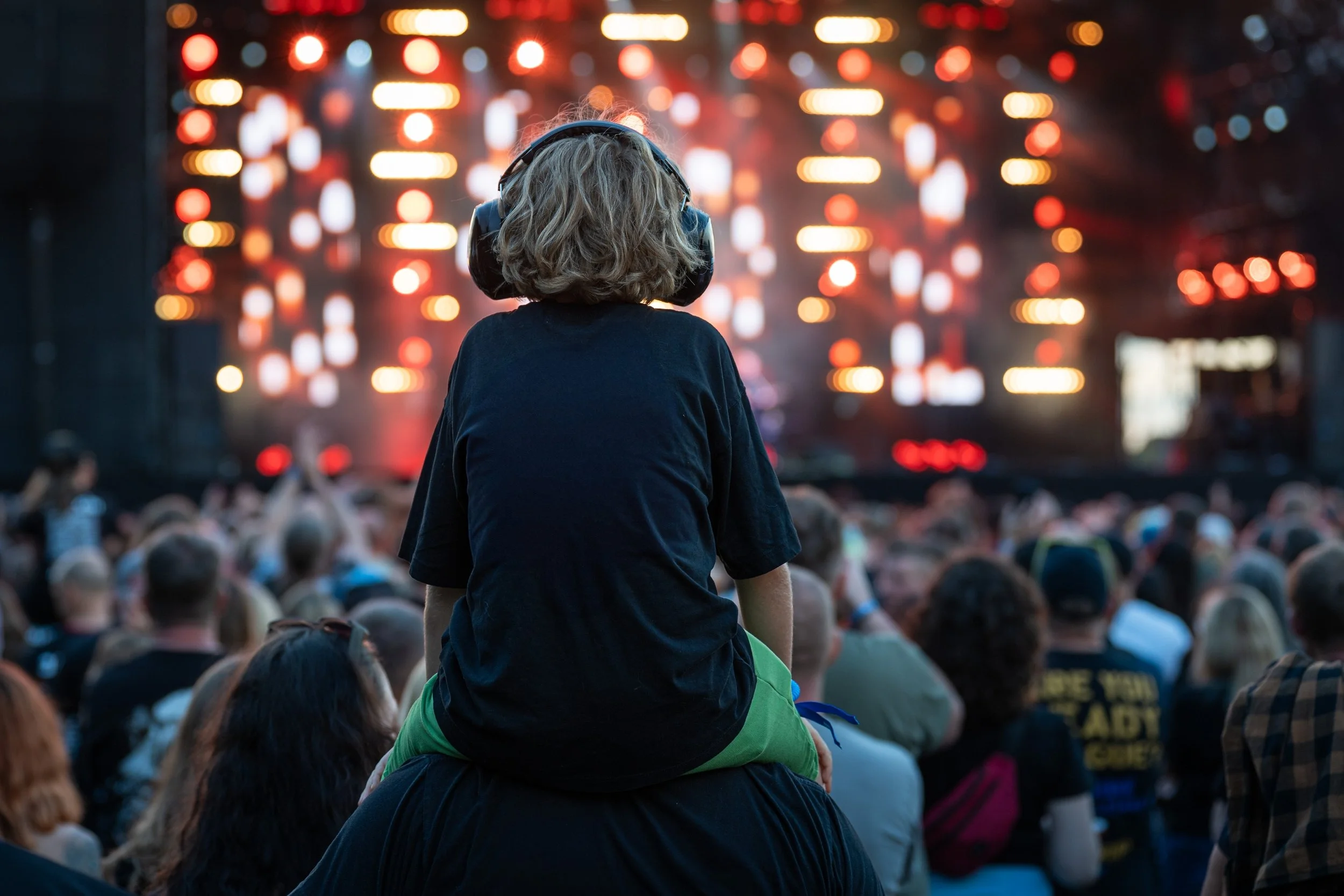 IRL: A little boy with headphones sitting on shoulders during a concert in front of the stage.
