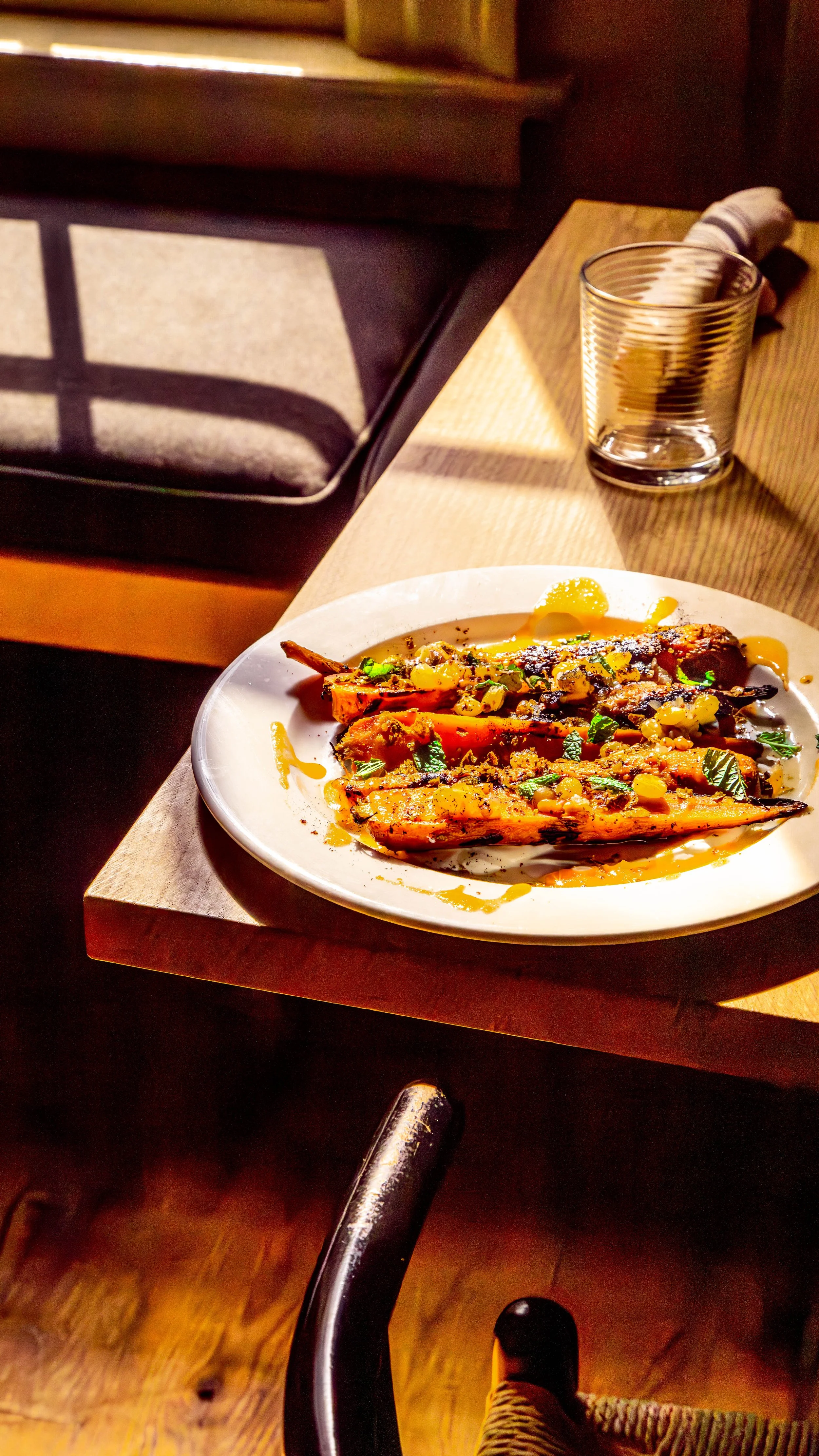 A white plate with grilled vegetables and sauce on a wooden table. A glass of water and a pair of salt and pepper shakers are nearby. Sunlight falls on the table.