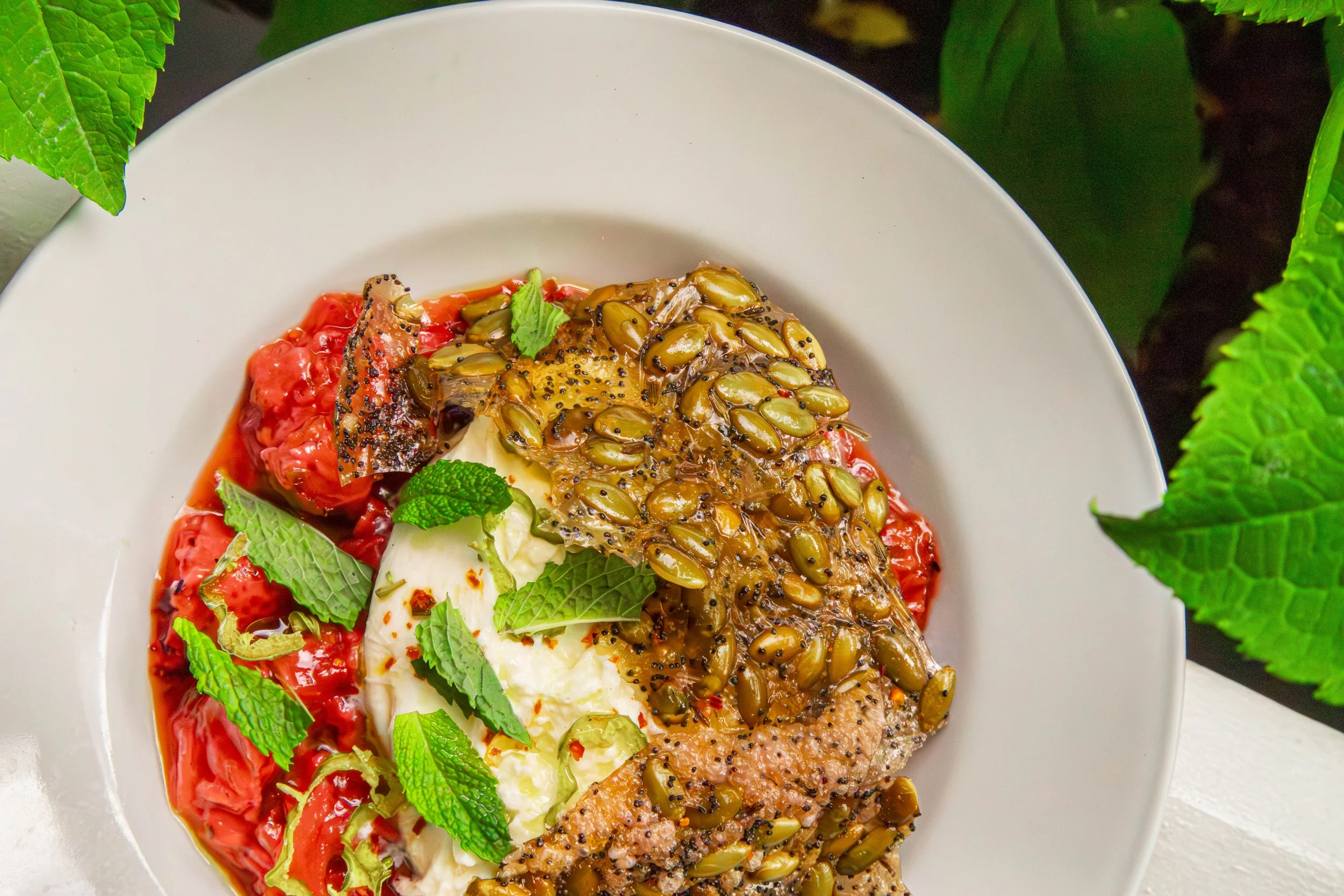 A plated dish featuring grilled fish topped with sunflower seed pesto, served with roasted red tomatoes, fresh mint leaves, and dollops of creamy mashed potatoes.