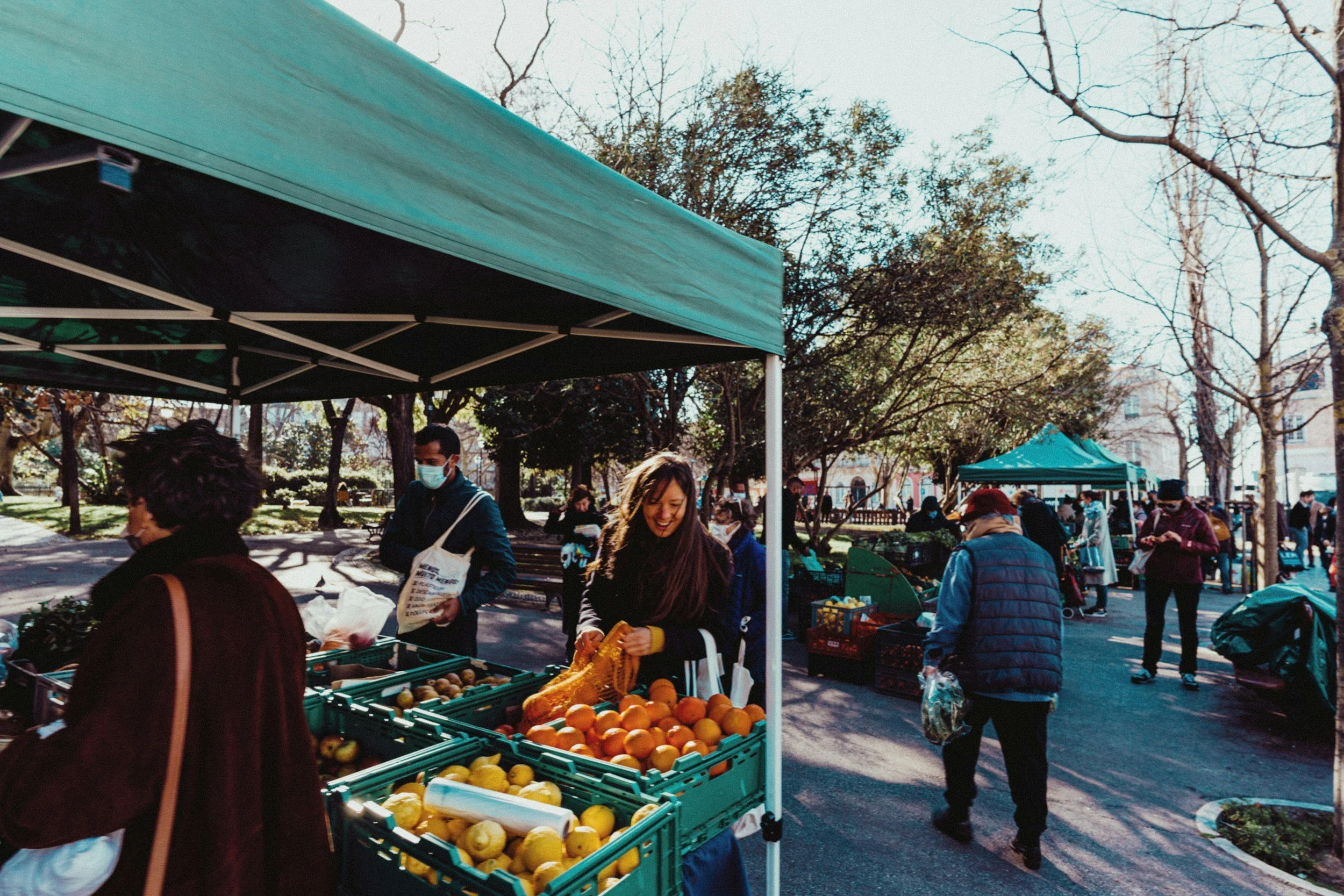 People shopping at an outdoor farmers market, browsing and purchasing fresh produce such as oranges, lemons, and other fruits, under a sunny sky with trees and additional market stalls in the background.