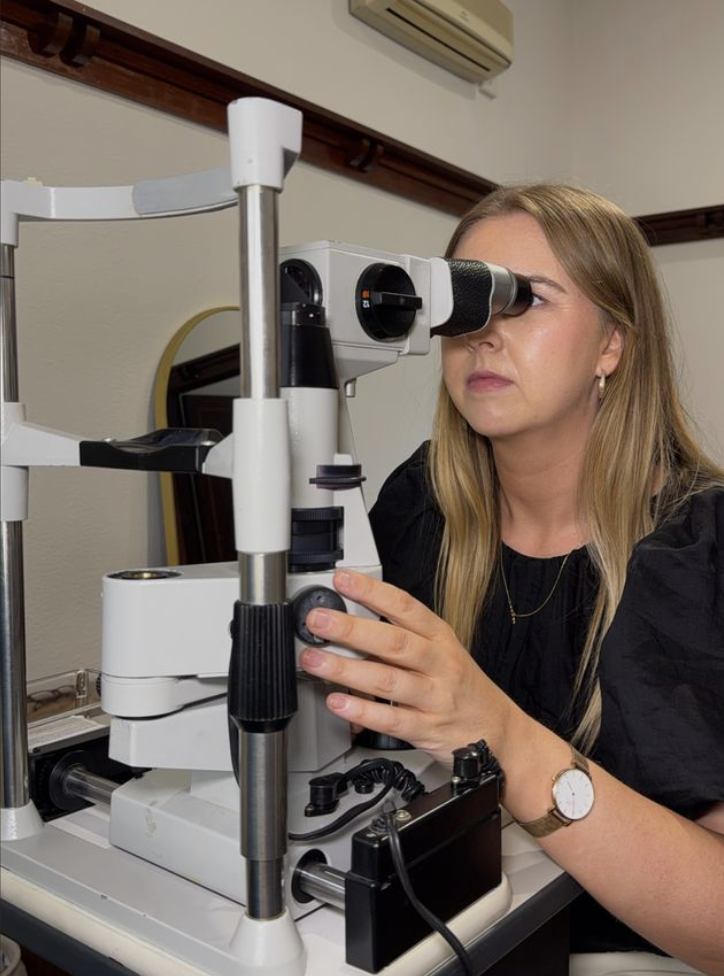 A woman with long blonde hair, wearing a black shirt and watch, using a microscope in a laboratory setting.