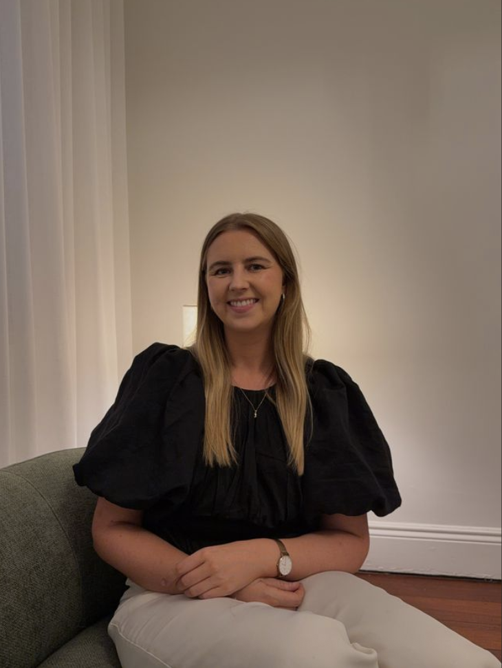 A young woman with long blonde hair is sitting on a green couch in a well-lit room, smiling at the camera. She is wearing a black top with puffed sleeves, white pants, a watch on her left wrist, and a necklace.