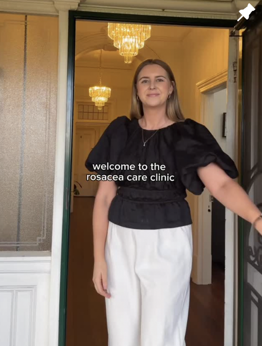 A woman standing at the entrance of a care clinic, wearing a black top and white pants, smiling. There is a chandelier hanging in the background inside the clinic.