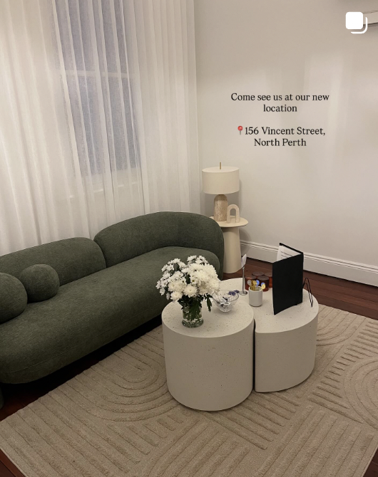 Living room with a dark green sofa, white curtains, and a beige rug. There is a small white side table with a lamp and decorative items, and two white rounded coffee tables with flowers, a bowl, and a standee on top. A wall sign invites to visit a new location at 156 Vincent Street, North Perth.