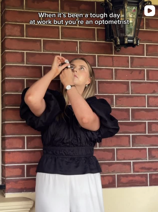 A woman in a black puff-sleeve blouse and white pants applying eye drops against a brick wall, with the caption 'When it's been a tough day at work but you're an optometrist'.