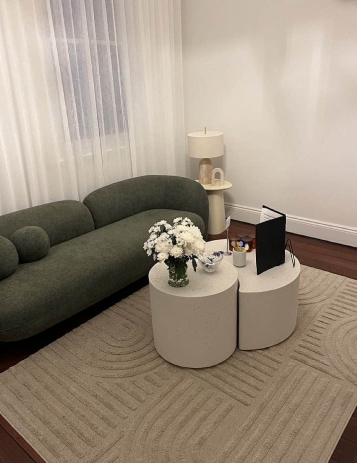 Living room with a green sofa, a beige lamp on a side table, white sheer curtains, and two white rounded coffee tables with flowers, a glass bowl, and other small items.