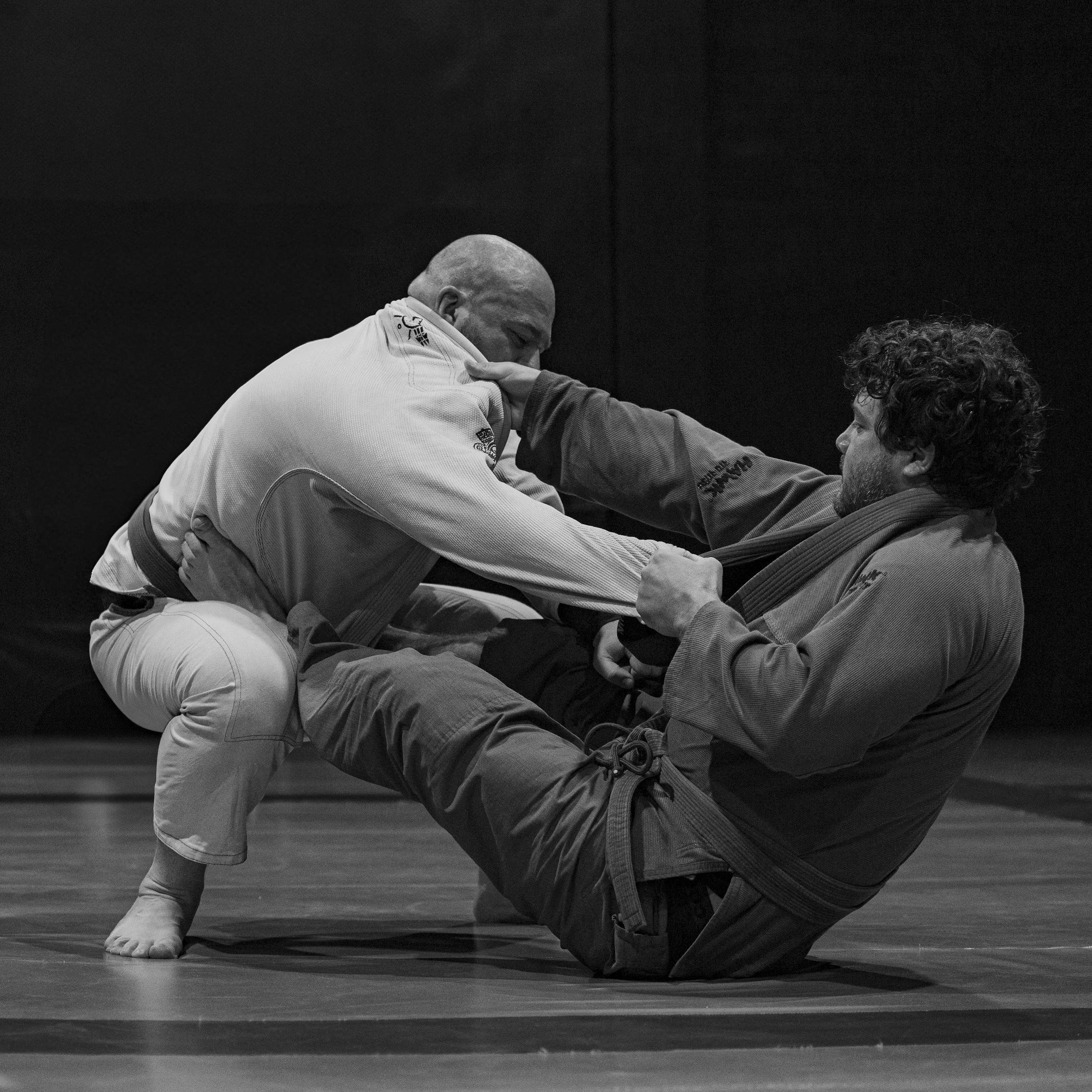 Two men practicing Brazilian Jiu-Jitsu on a mat, one in a white gi and the other in a dark gi, engaged in a grappling exchange.