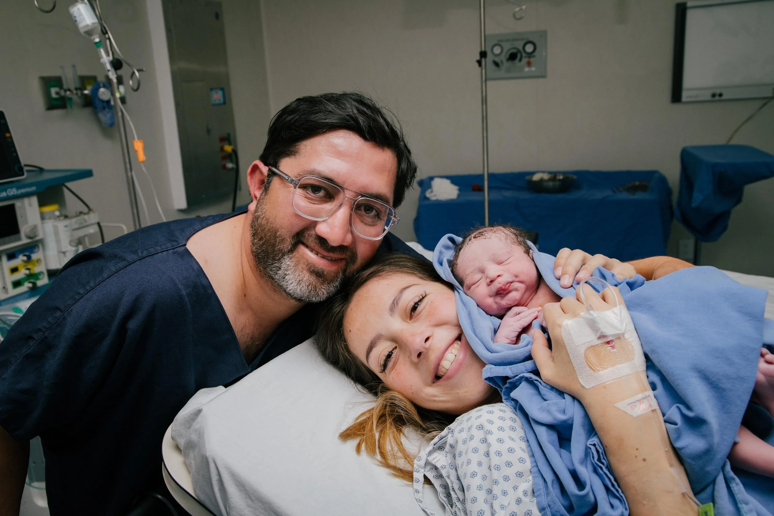 Una mujer recién dada a luz en un hospital, con un hombre y un bebé en brazos, todos sonriendo. documentando el parto de manera humana y amorosa
