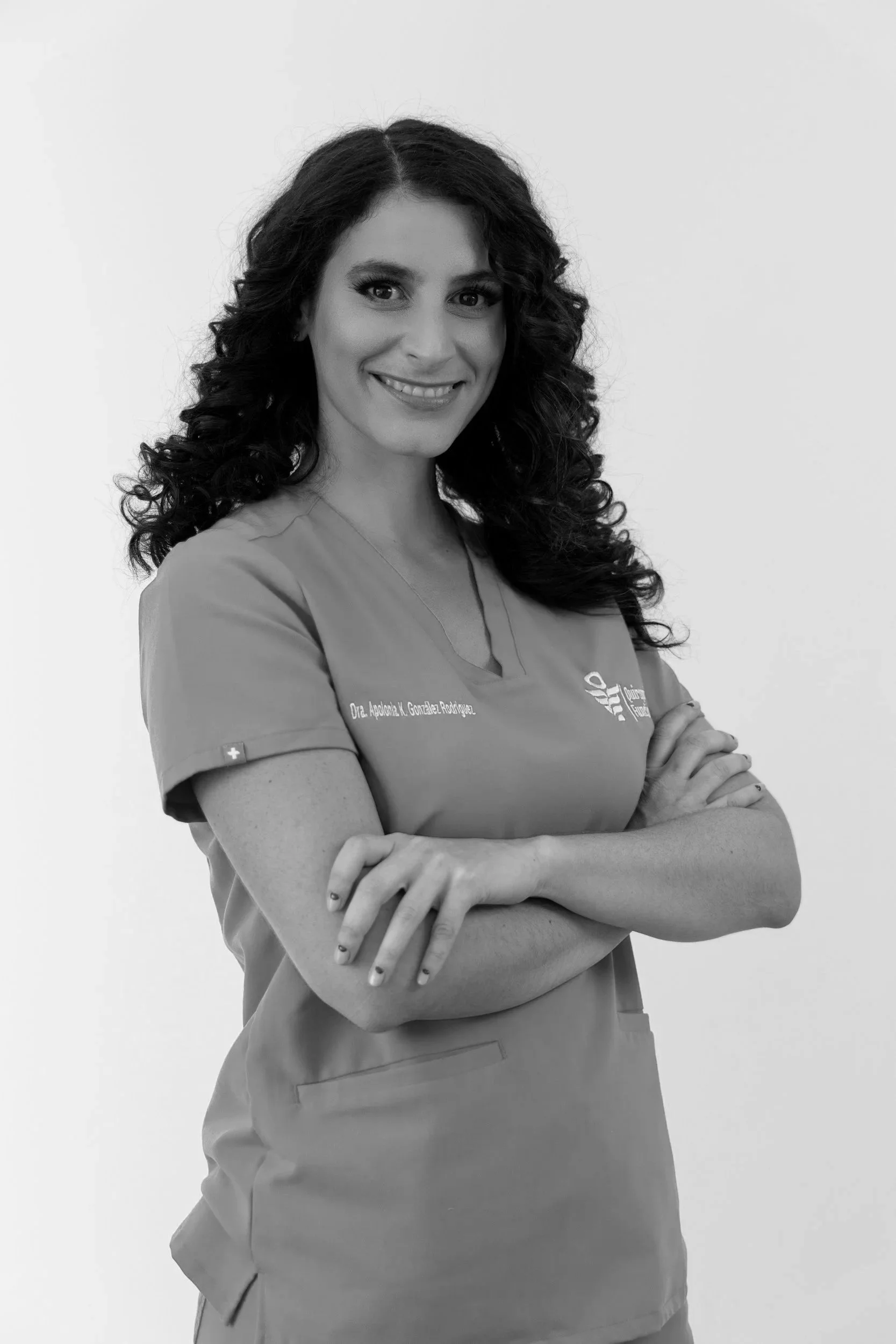 Mujer sonriente con cabello rizado y uniforme de enfermera, con brazos cruzados, en un fondo blanco.
