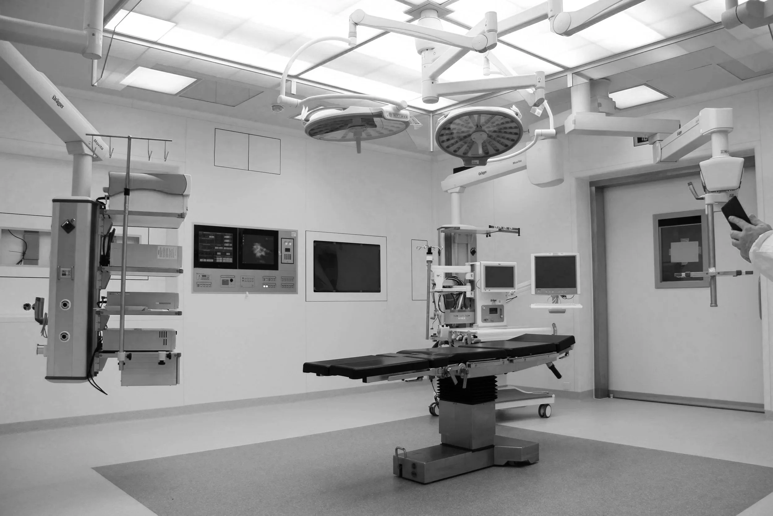 Sala de cirugía moderna con equipos médicos, monitores y lámparas quirúrgicas, en blanco y negro.
